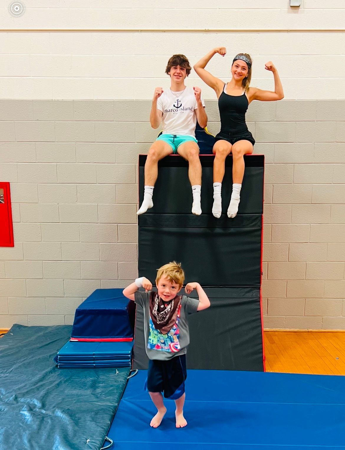 A boy is flexing his muscles in front of two women sitting on blocks in a gym.