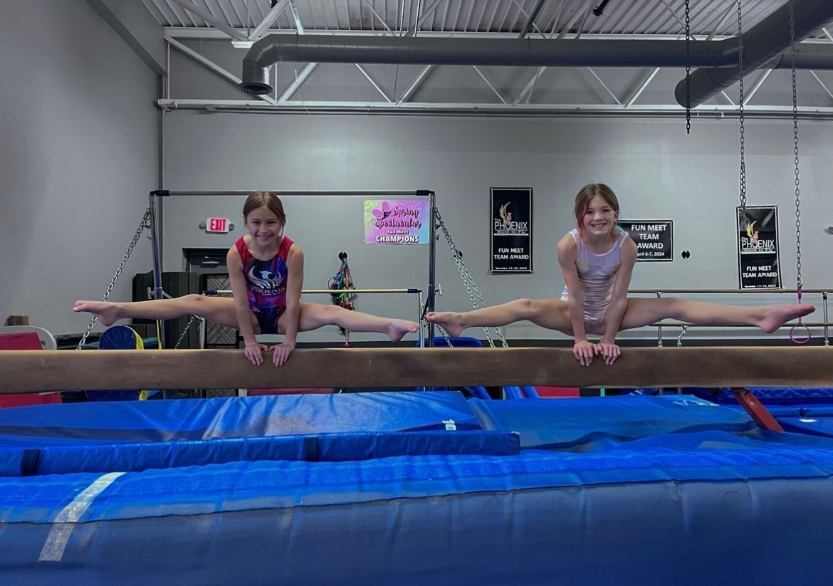 Two young girls are doing splits on a balance beam in a gym.