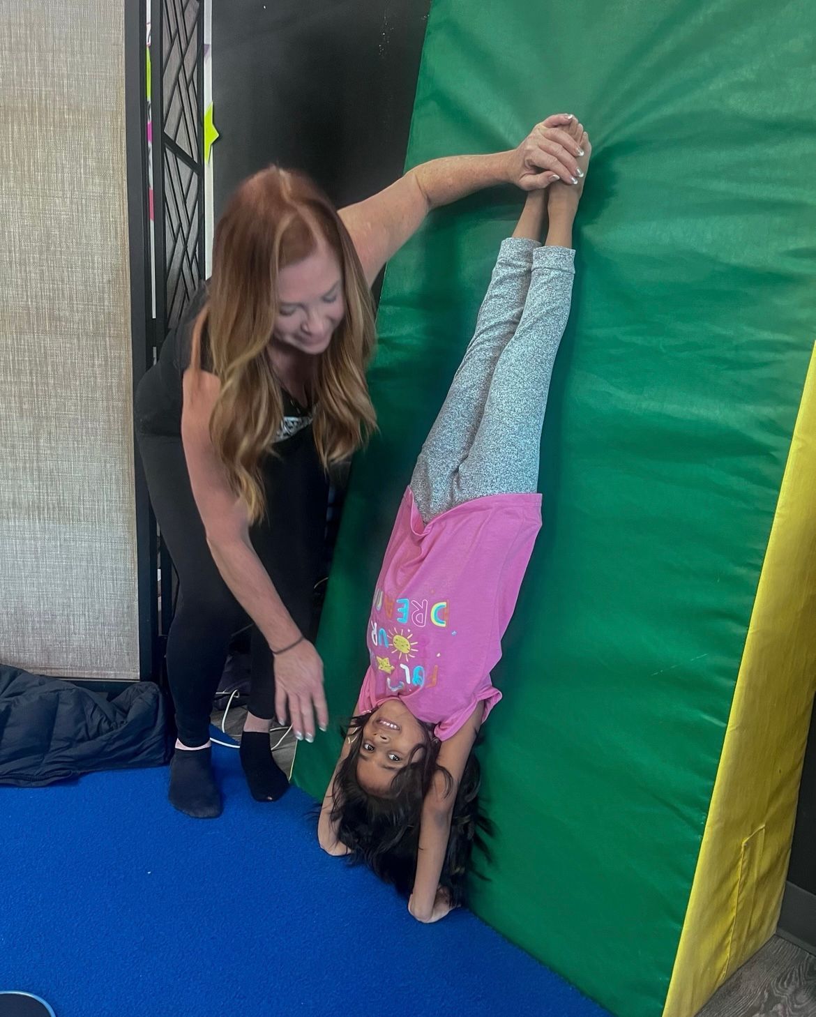 A woman is helping a little girl do a handstand on a green mat.