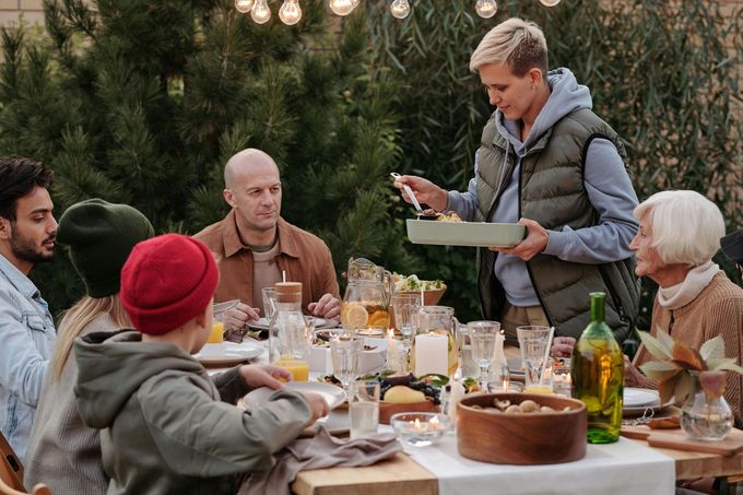 Family gathering outdoors, person serving food at a wooden table, festive lights, smiling faces.