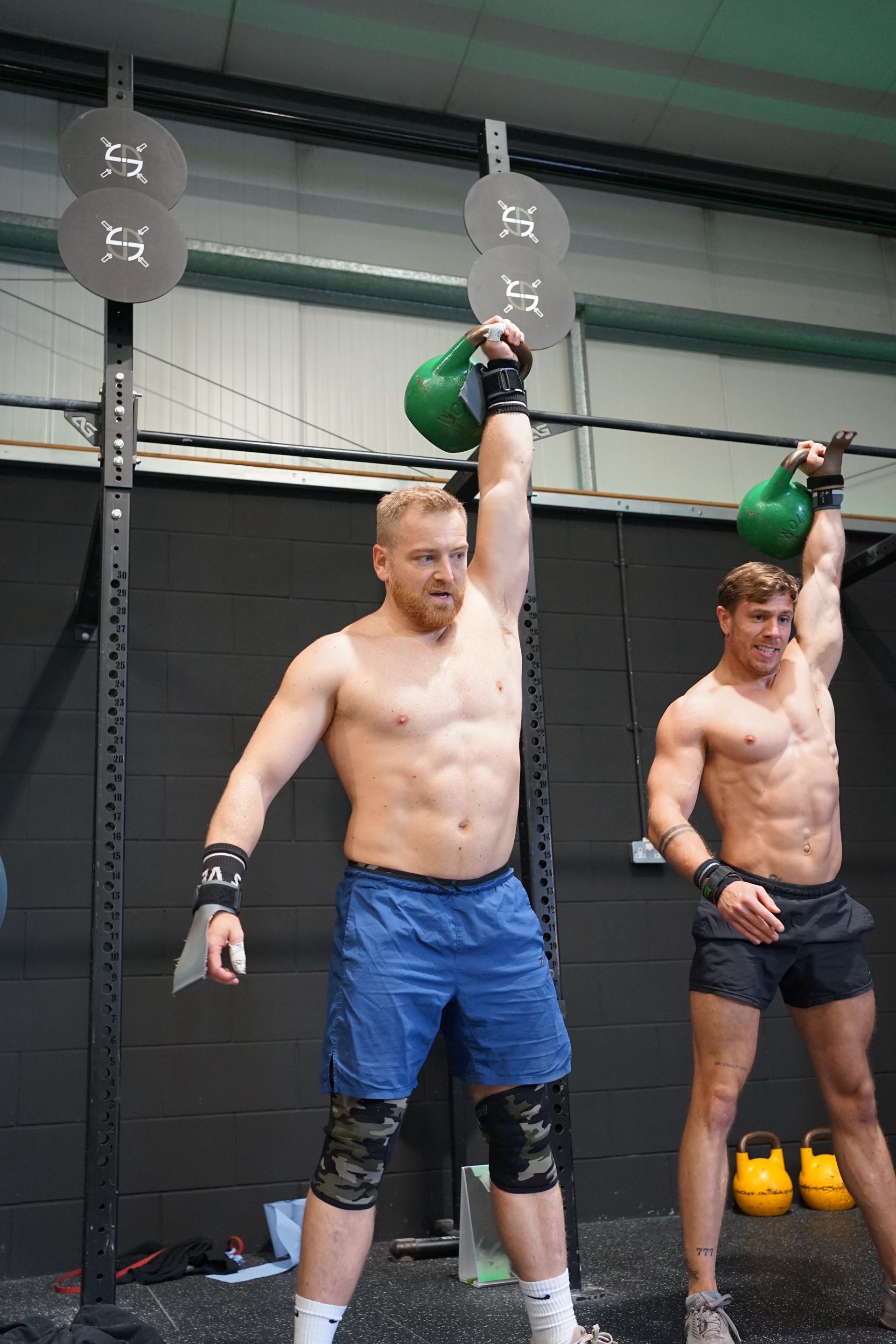 Two shirtless men are lifting kettlebells in a gym.