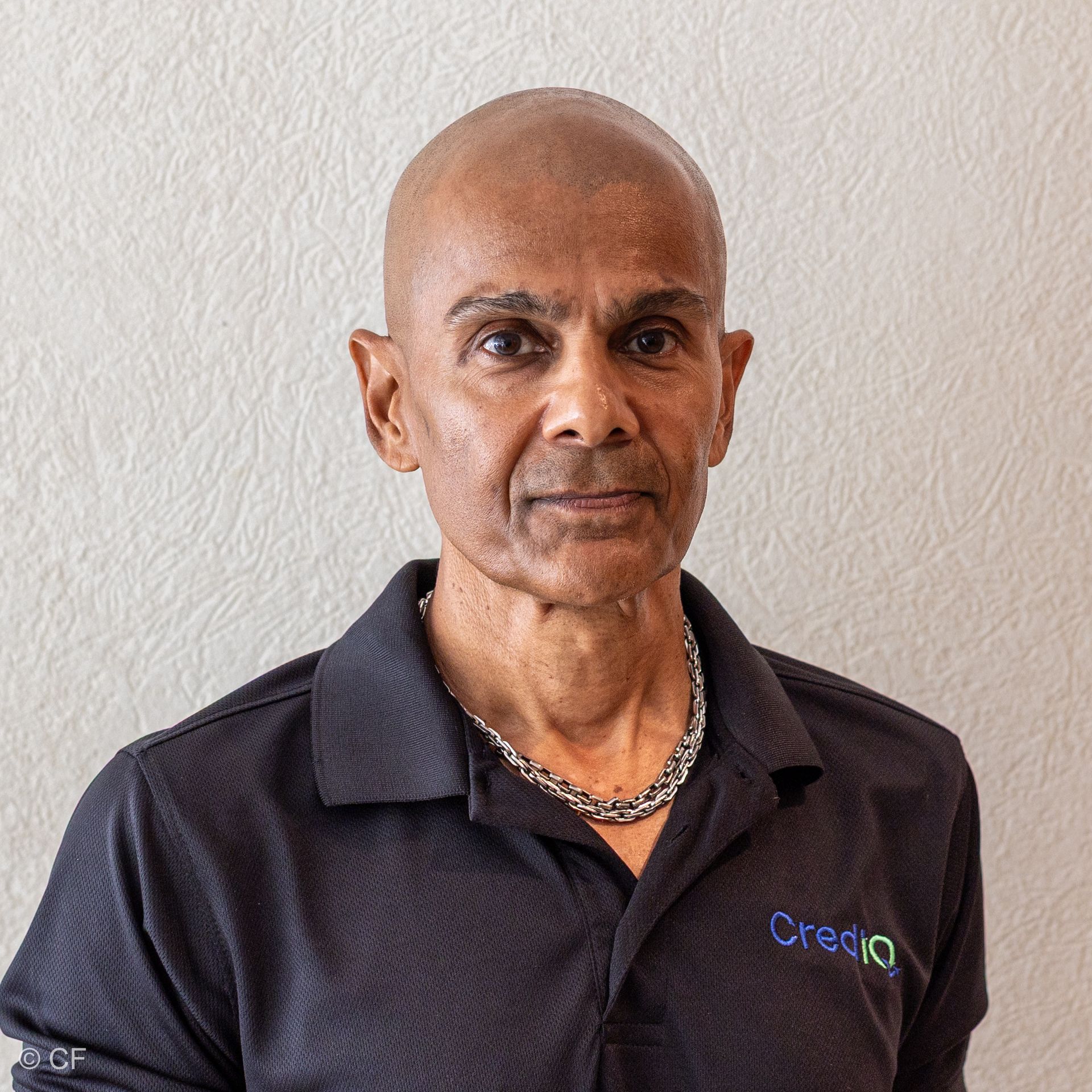 Razeem Ali wearing a dark polo shirt with a chain necklace, posed against a light, textured background.