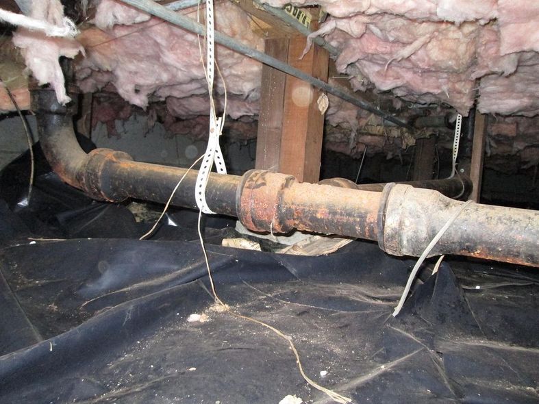 Rusty pipe running through a crawlspace under exposed insulation and joists