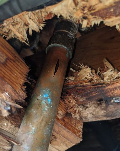 Rusty copper pipe inside a damaged wooden structure with frayed insulation and blue paint specks