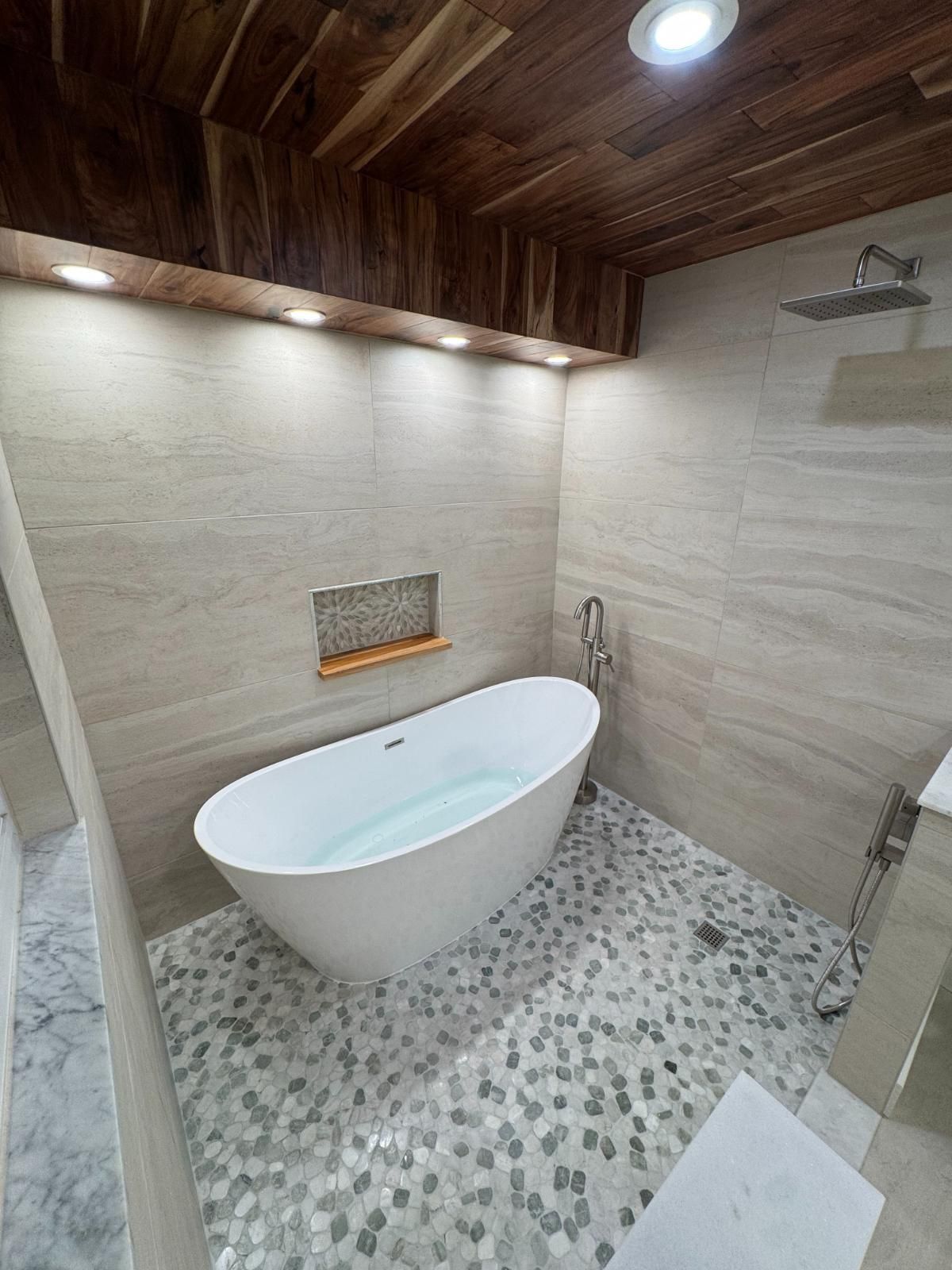 Modern bathroom with freestanding white tub, stone-tile walls, pebble floor, and wood ceiling accents