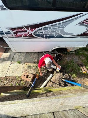 Person in red shirt kneeling by a white trailer, working on a muddy ground area with tools.