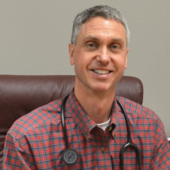 Dr. Noah wearing a plaid shirt and stethoscope smiles in a medical office setting.