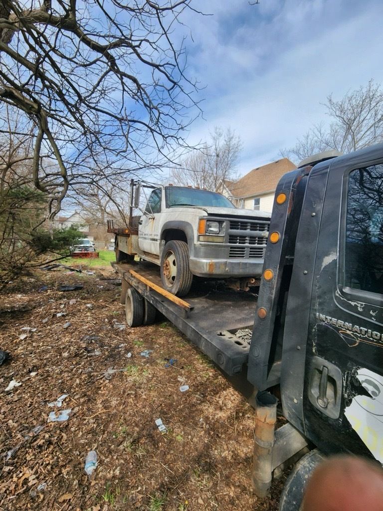 Tow truck hauling a white pickup truck on a dirt road, trees, and a house in the background.