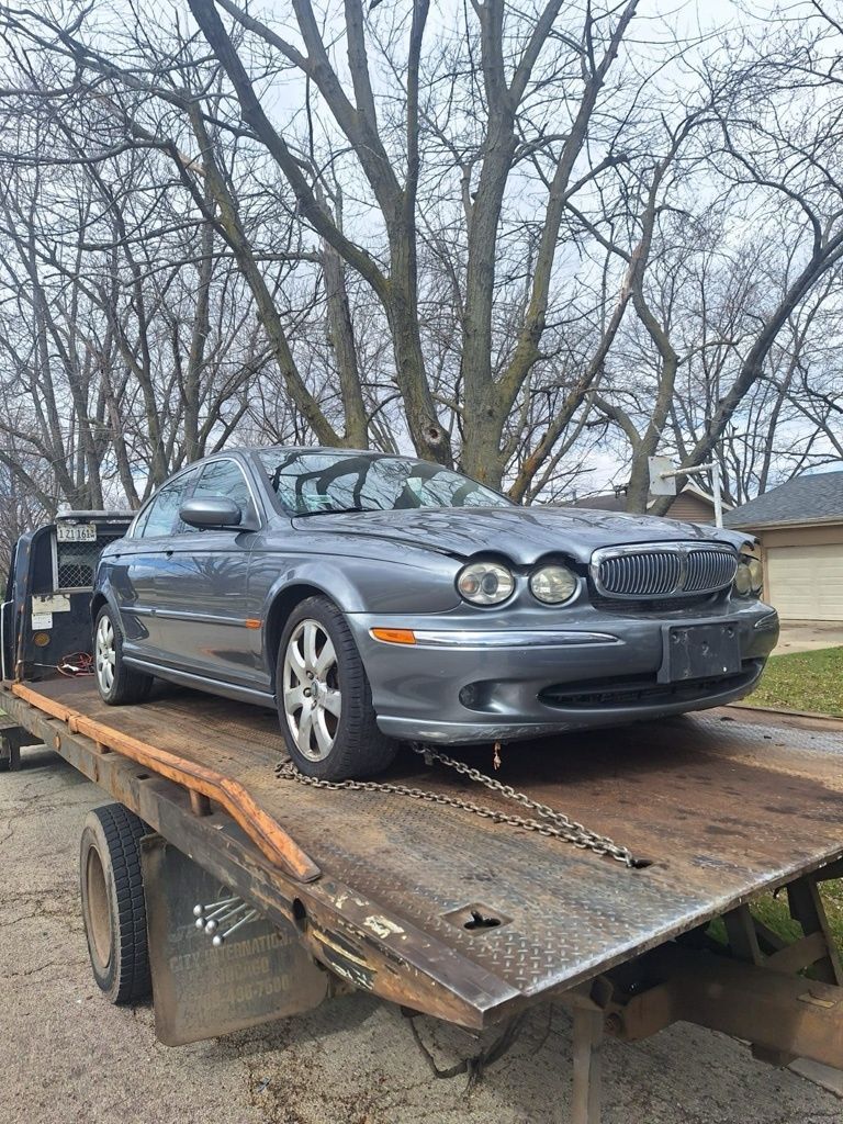 Gray Jaguar X-Type sedan on a flatbed tow truck, likely being transported. Outdoors, overcast sky.