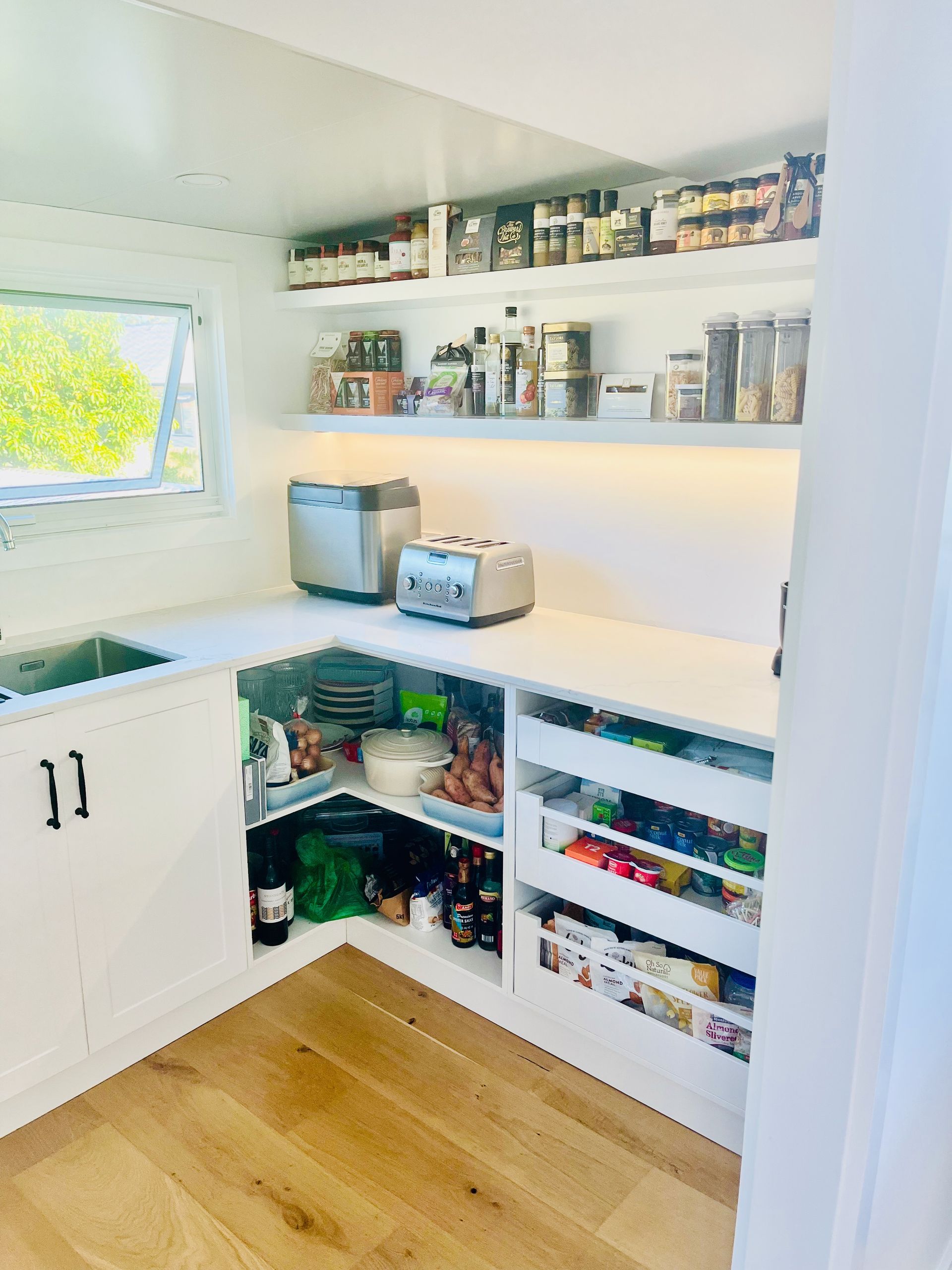 Small white pantry with shelves filled with food and spices, countertop with toaster and rice cooker, and a window.