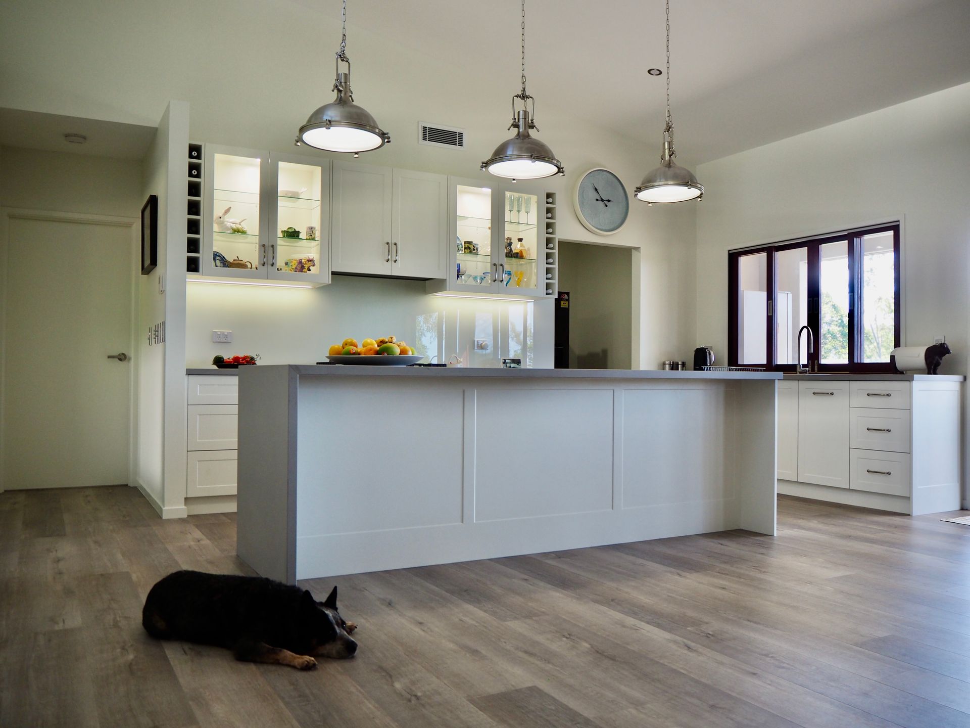 Kitchen With Island, Pendant Lights, And A Dog Lying On The Wooden Floor. White Cabinets And Countertops, Light Gray Floor