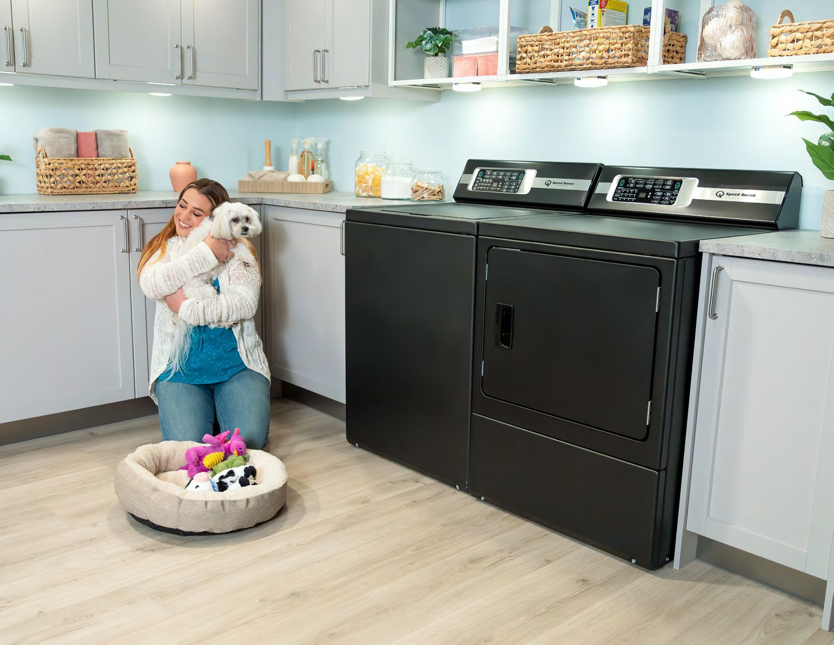 A woman is kneeling down in a laundry room holding a small white dog.