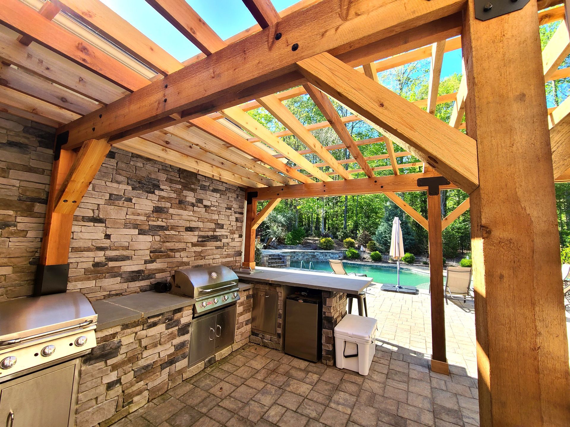An outdoor kitchen with stone counters and stainless appliances under a rustic timber pergola, overlooking a pool.