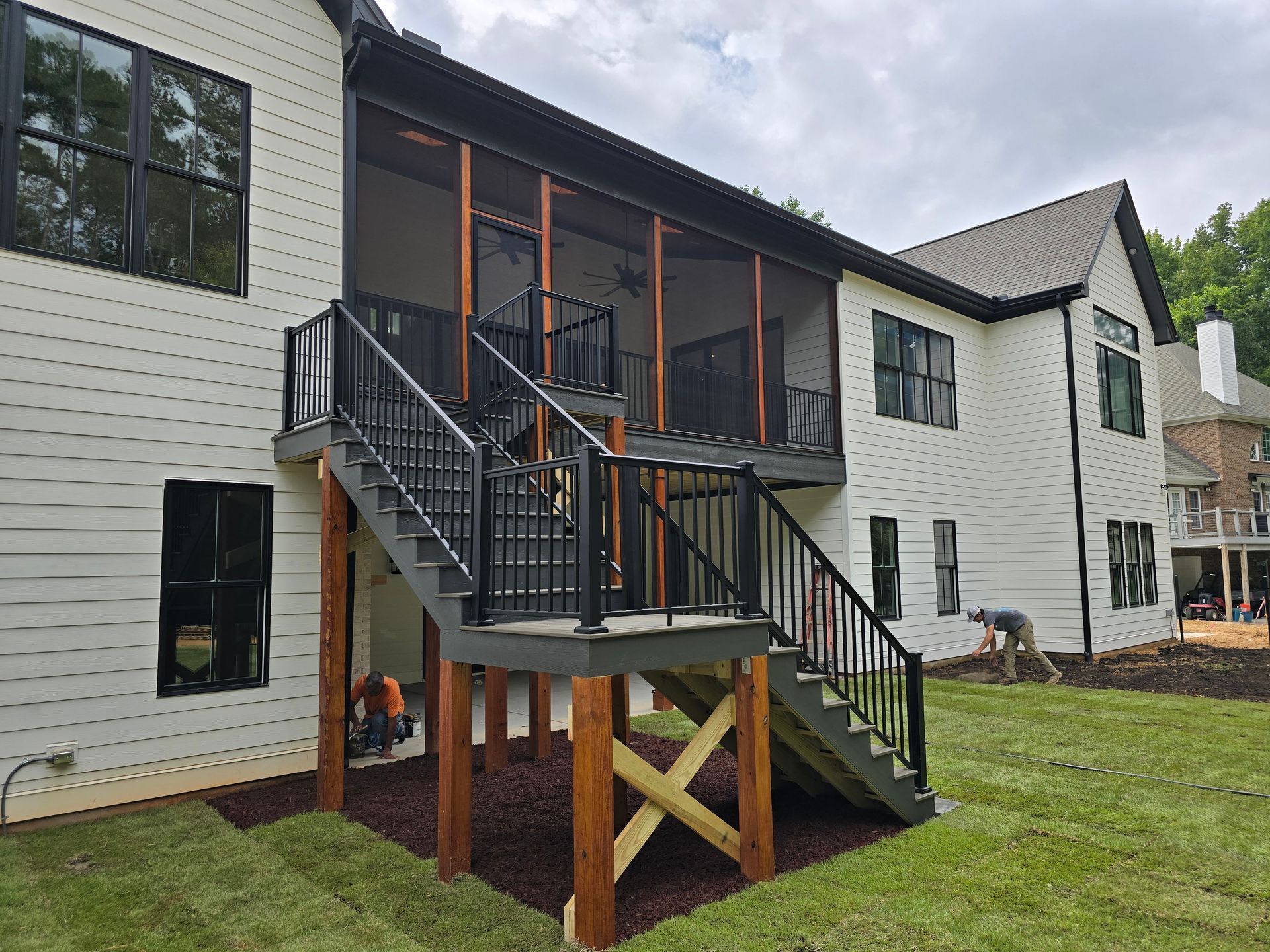 Two workers construct a wooden deck and staircase attached to the back of a light-colored house with a screened-in porch.