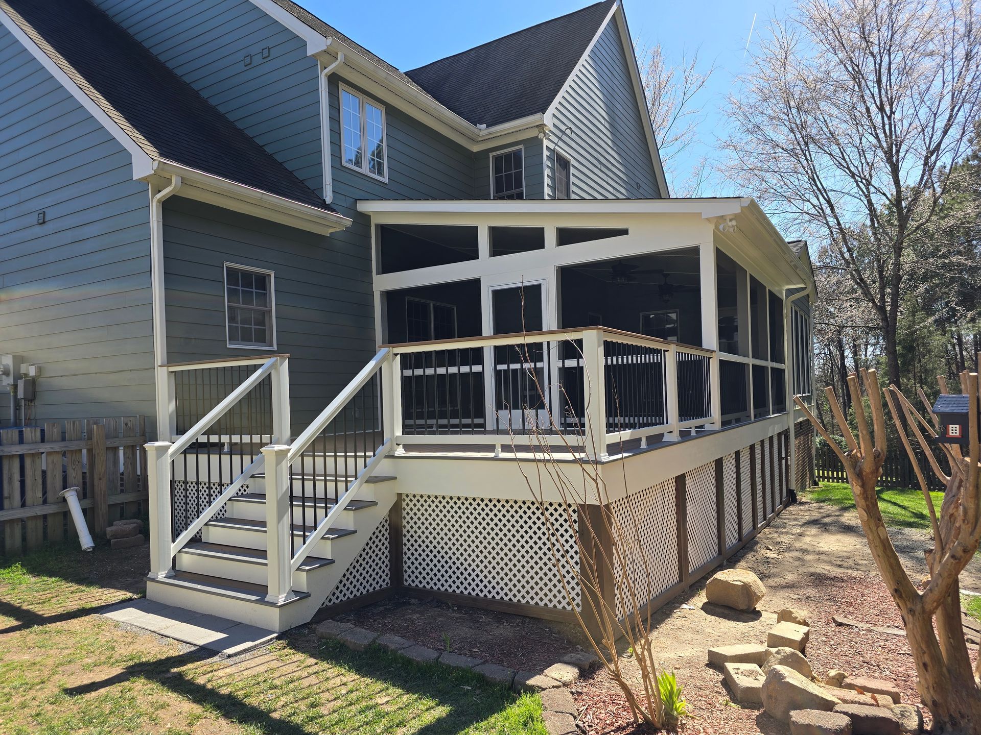 A gray house with a white screened-in porch, a wooden deck, and white stairs leading down to a lawn on a sunny day.