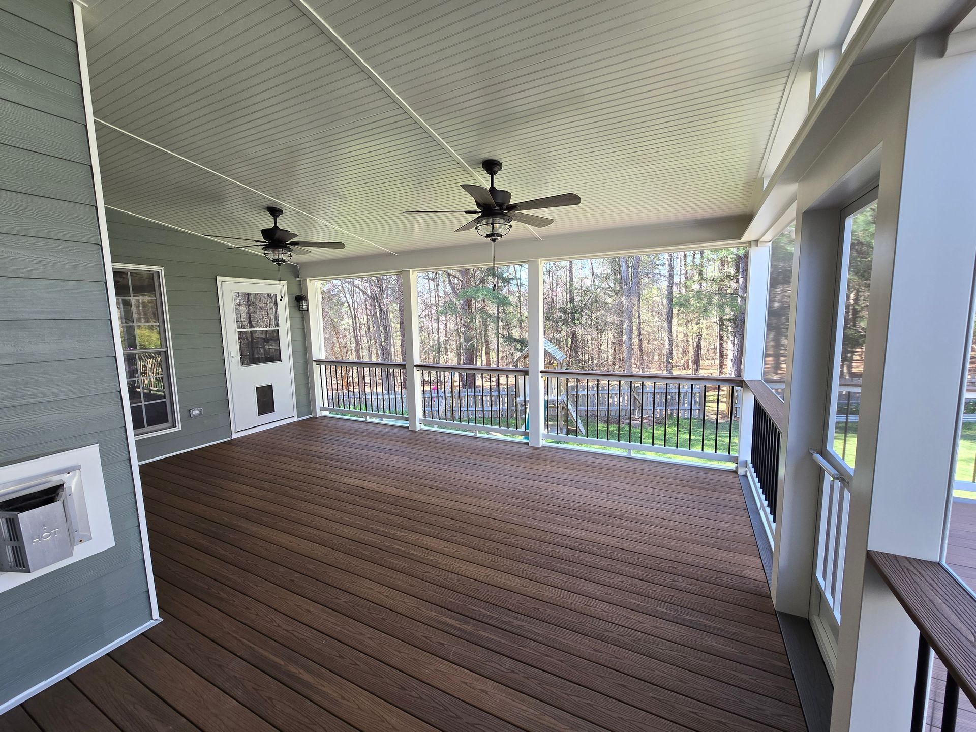 A spacious, covered outdoor porch with dark brown decking, grey siding, white trim, and two ceiling fans.