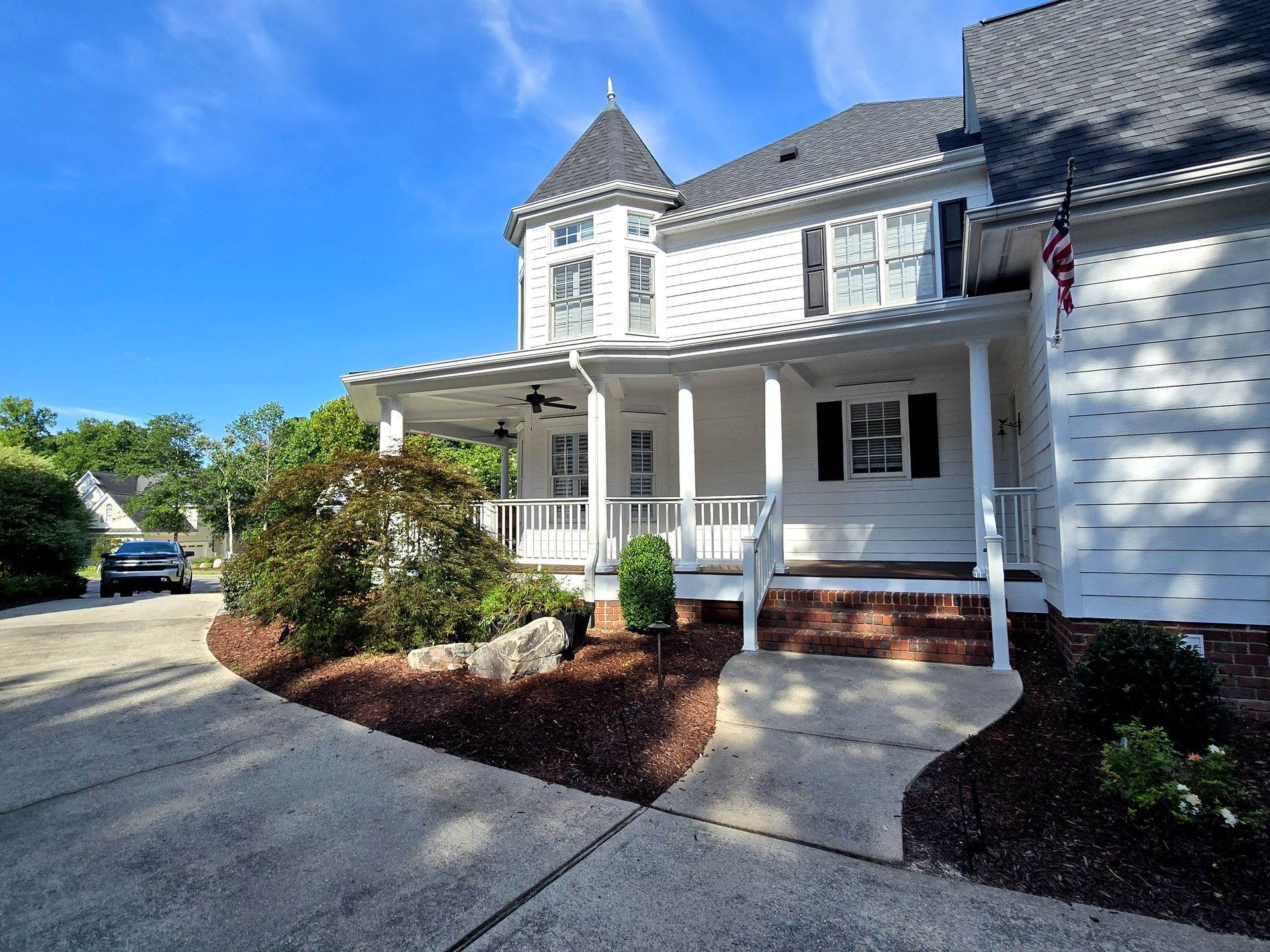 A white two-story house with a turret, covered front porch, and brick steps, situated by a paved driveway.