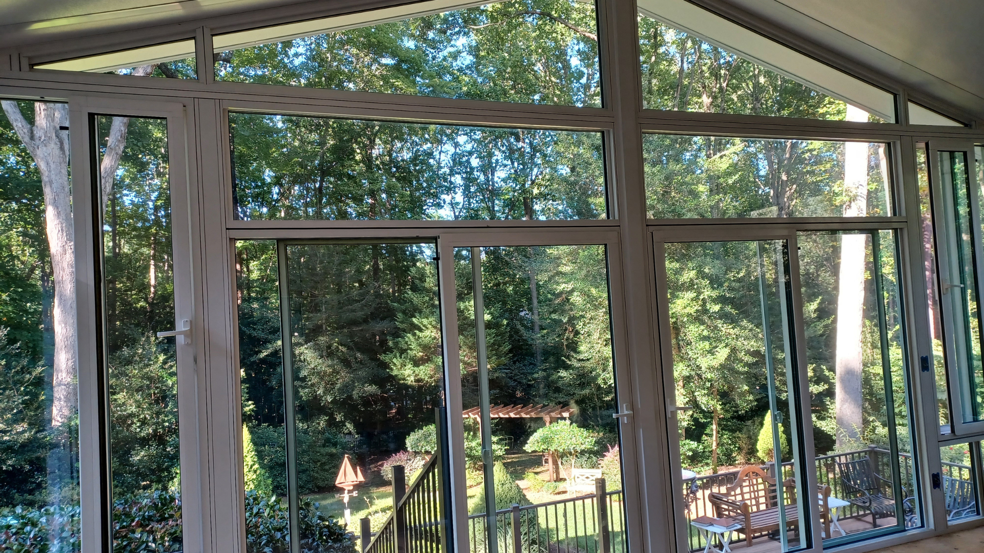 A view from inside a sunroom through large windows looking out at a lush, green wooded yard on a sunny day.