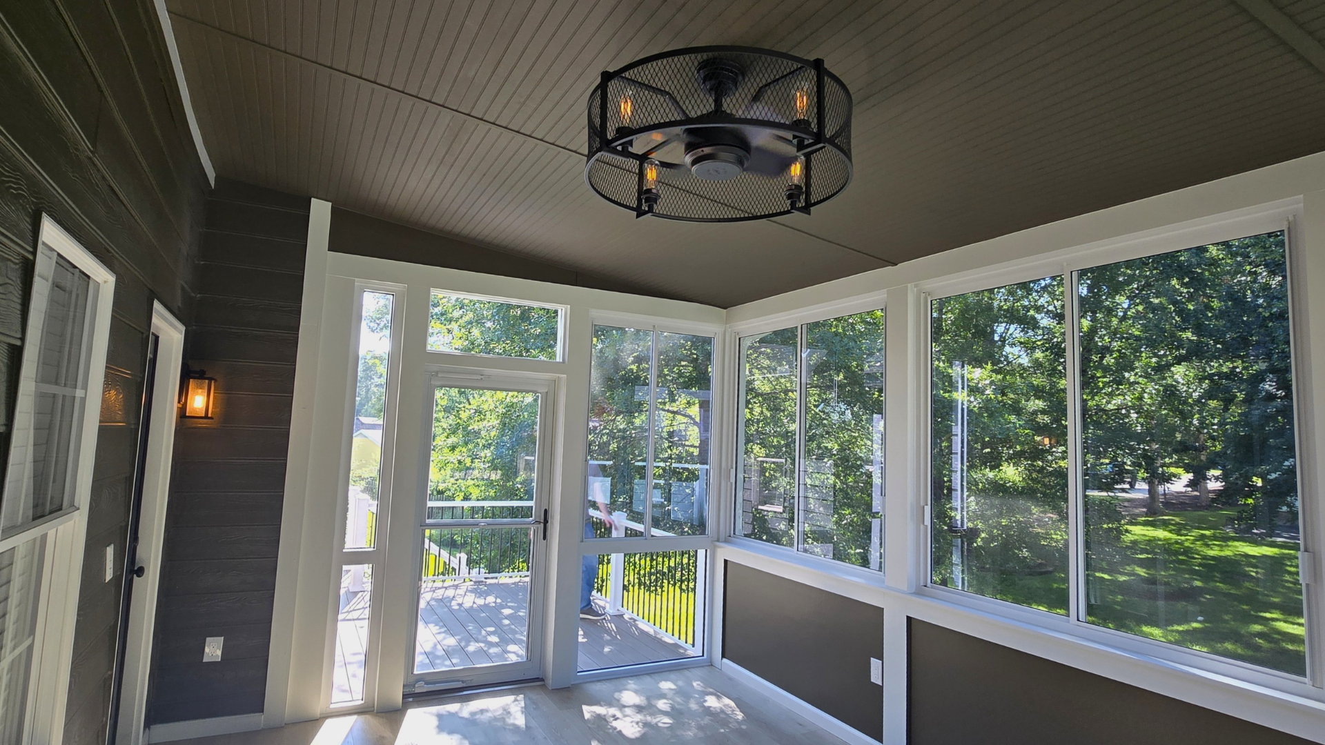 A sunroom with dark gray walls, a wooden ceiling, a cage-style ceiling fan, and large windows looking out onto trees.