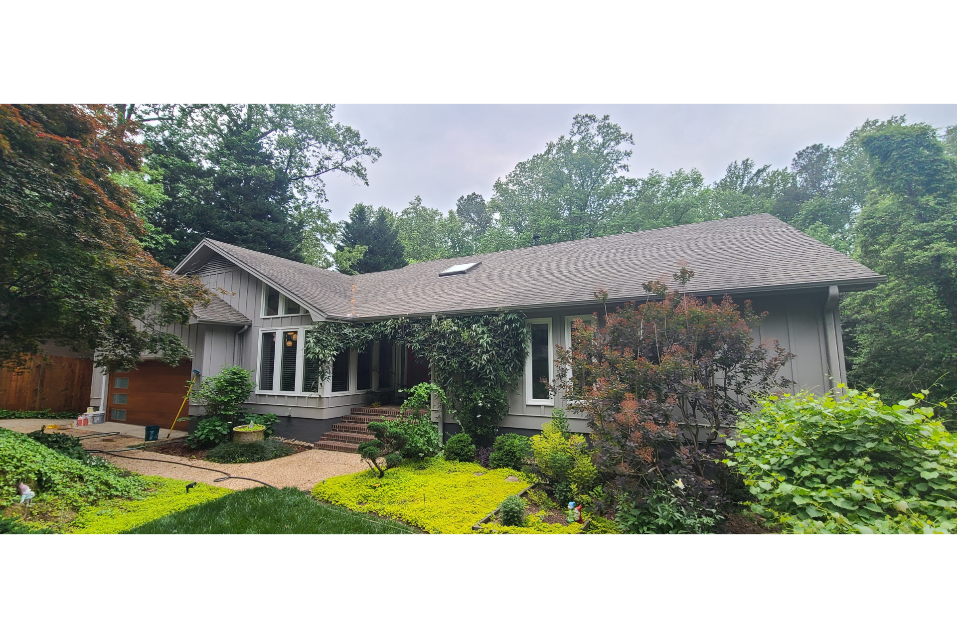 A single-story gray house with a brown shingled roof, surrounded by green trees, lush landscaping, and a gravel driveway.
