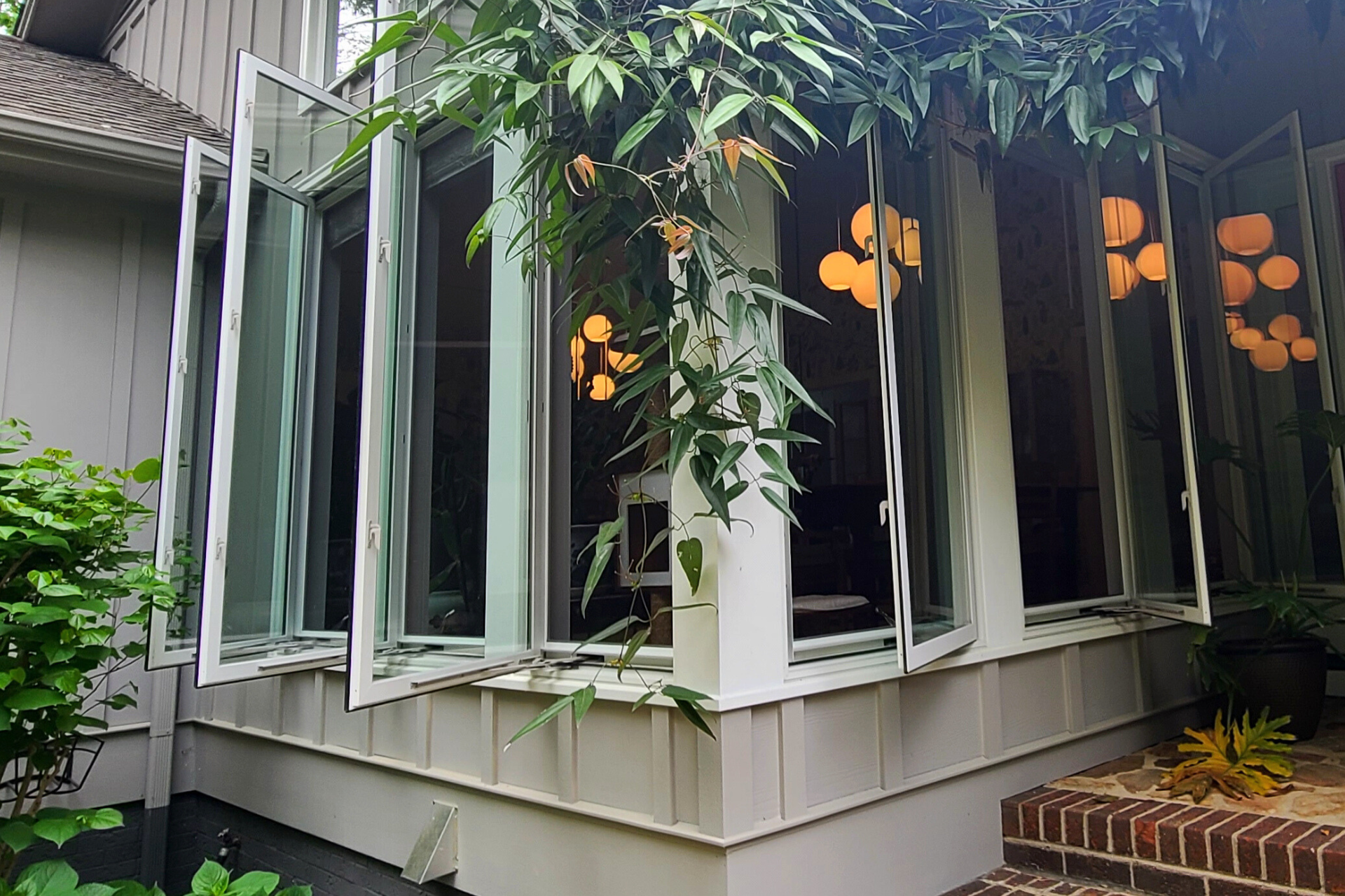 An exterior view of a cream-colored sunroom with large, open casement windows and hanging globe lights visible inside.