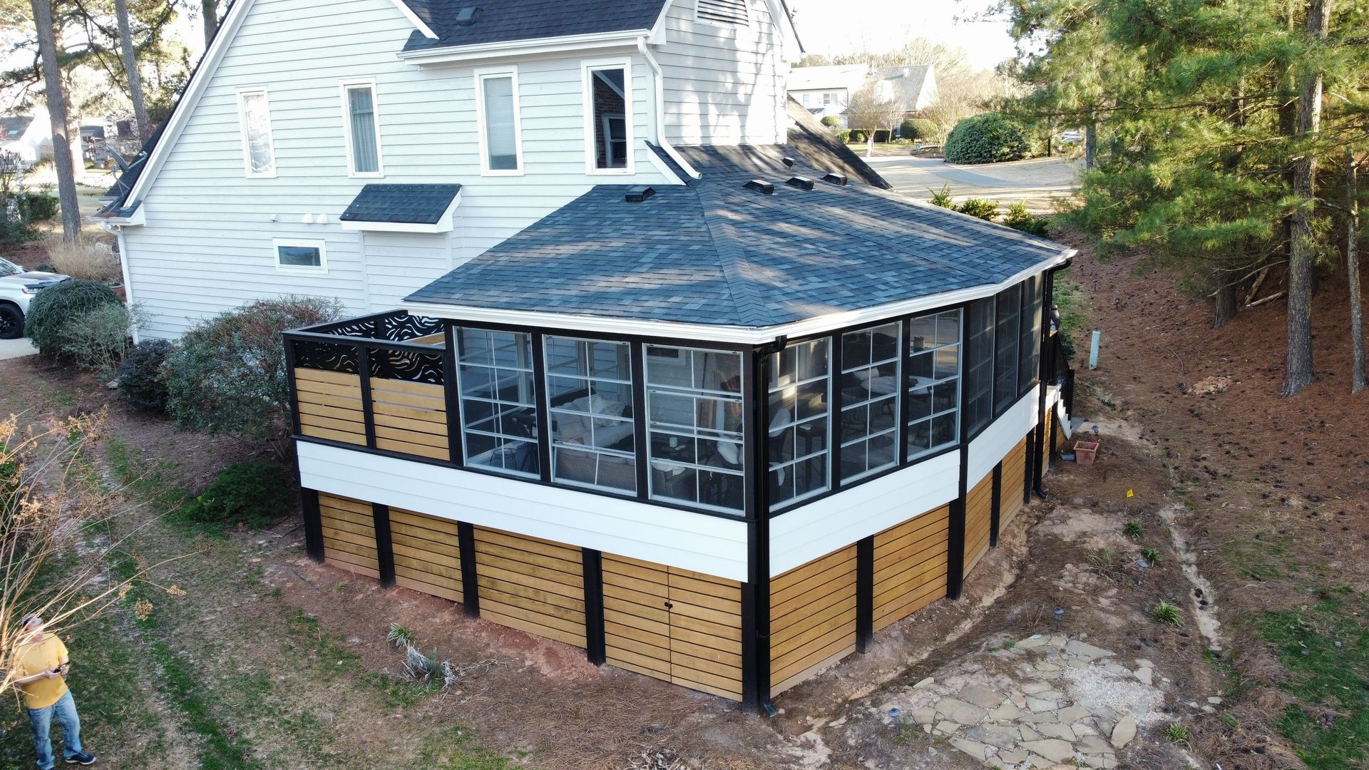 An elevated sunroom addition with glass windows, a dark shingled roof, and horizontal wood lattice skirting on a white house.