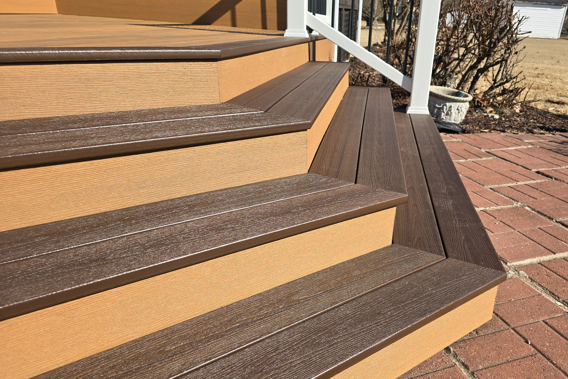 A set of multi-level outdoor composite stairs with brown wood-grain treads and tan risers, leading onto a brick patio.