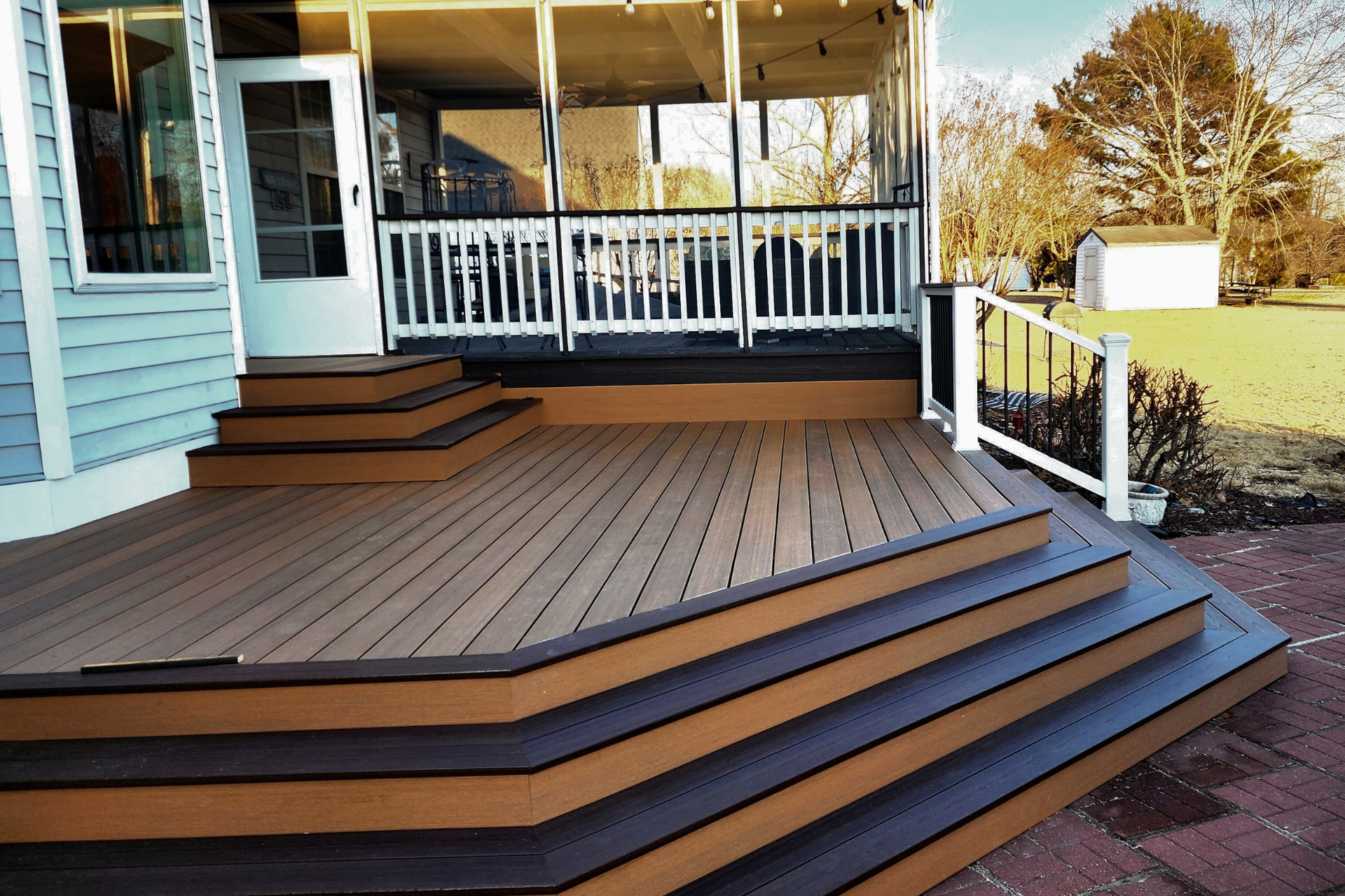 A multi-level outdoor deck featuring brown wood planks with dark trim, stairs, and a white railing leading to a porch.