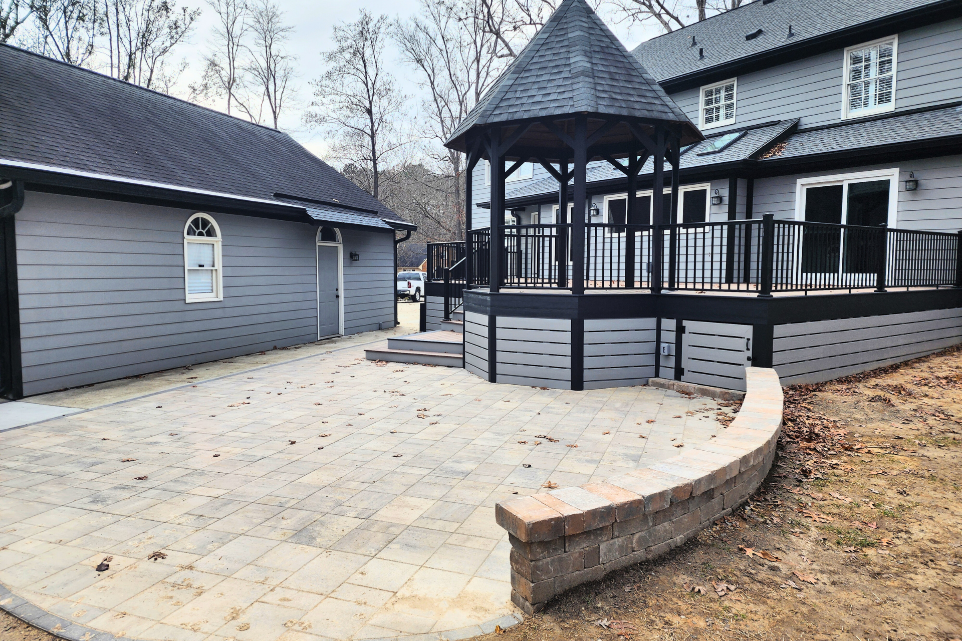 A gray house with a stone patio, a curved stone retaining wall, and a raised wooden deck with a black gazebo.