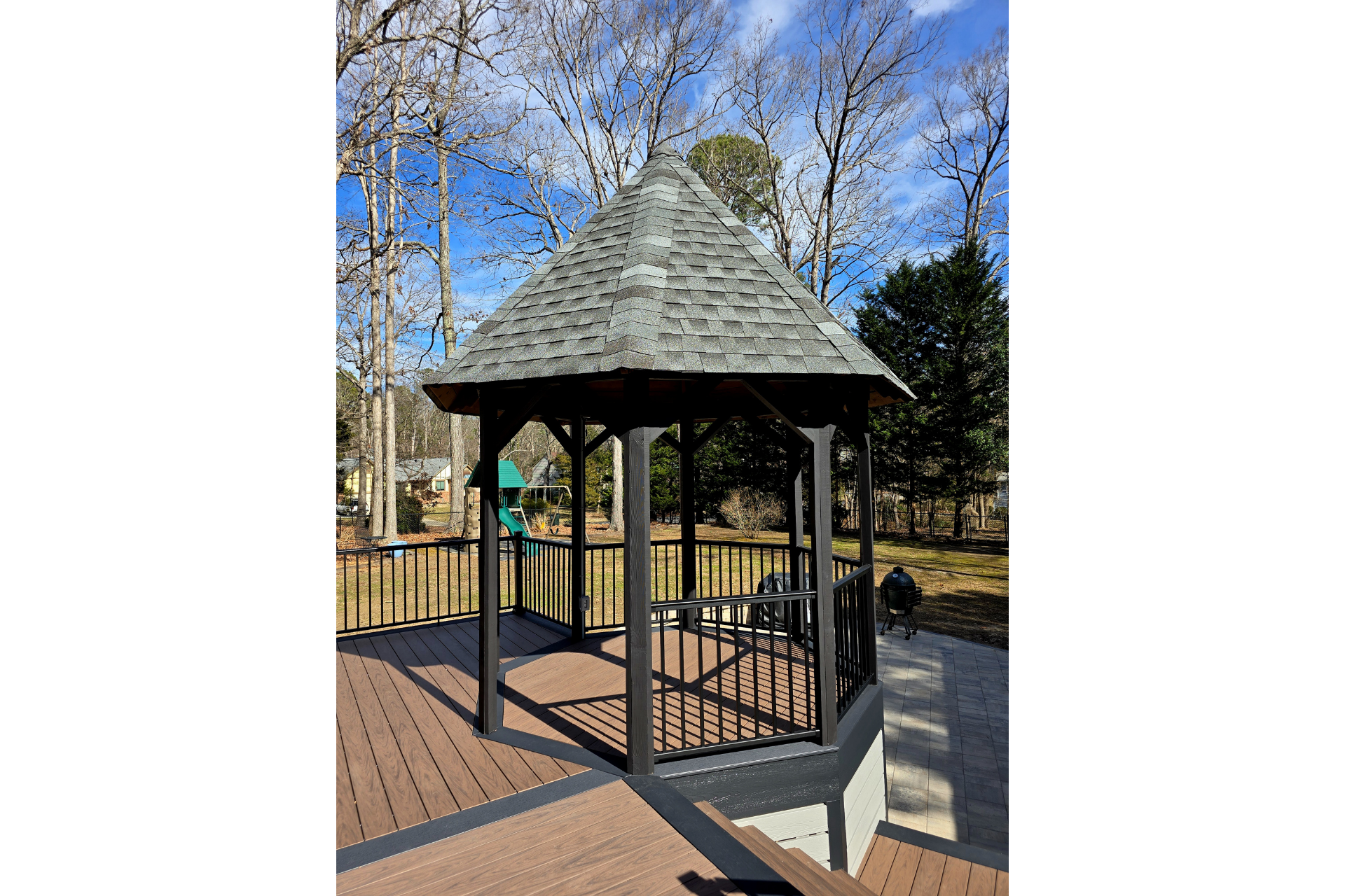 A black wooden gazebo with a shingled roof stands on a deck in a backyard with trees under a blue sky.