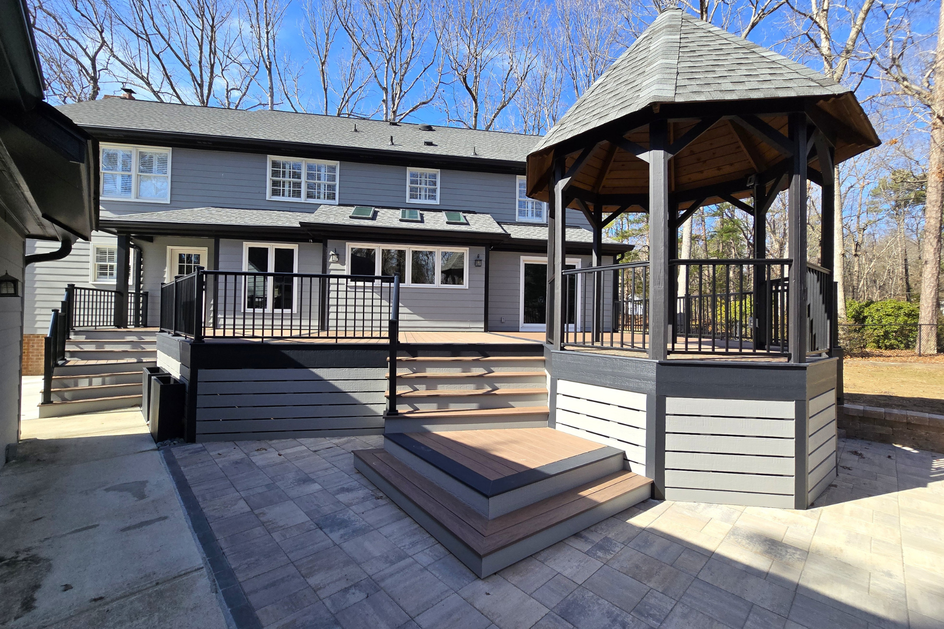 A gray two-story house with a wooden deck and a gazebo on a paved patio area.