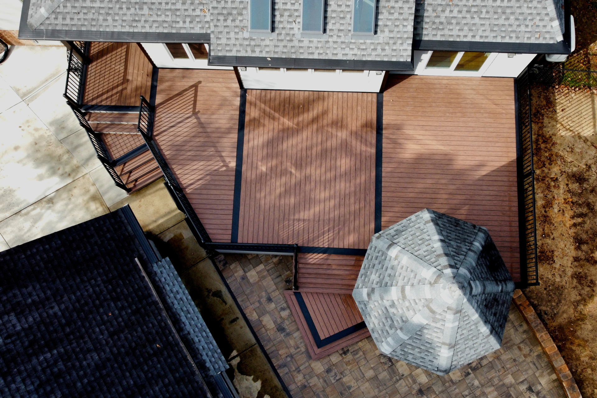 Aerial view of a tiered wooden deck with black railings and a matching pergola attached to the rear of a residential home.
