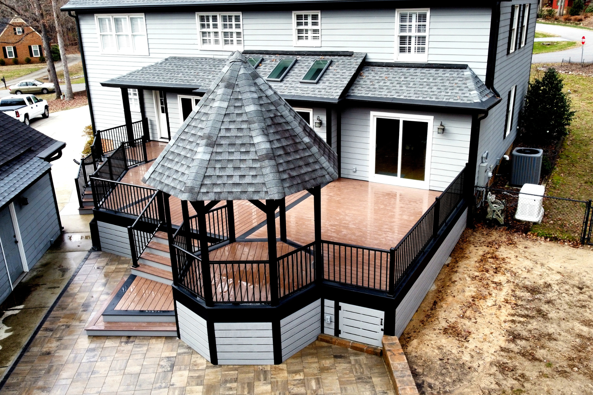 A high-angle view of a grey house with a multi-level deck, black railings, and a central gazebo overlooking a stone patio.