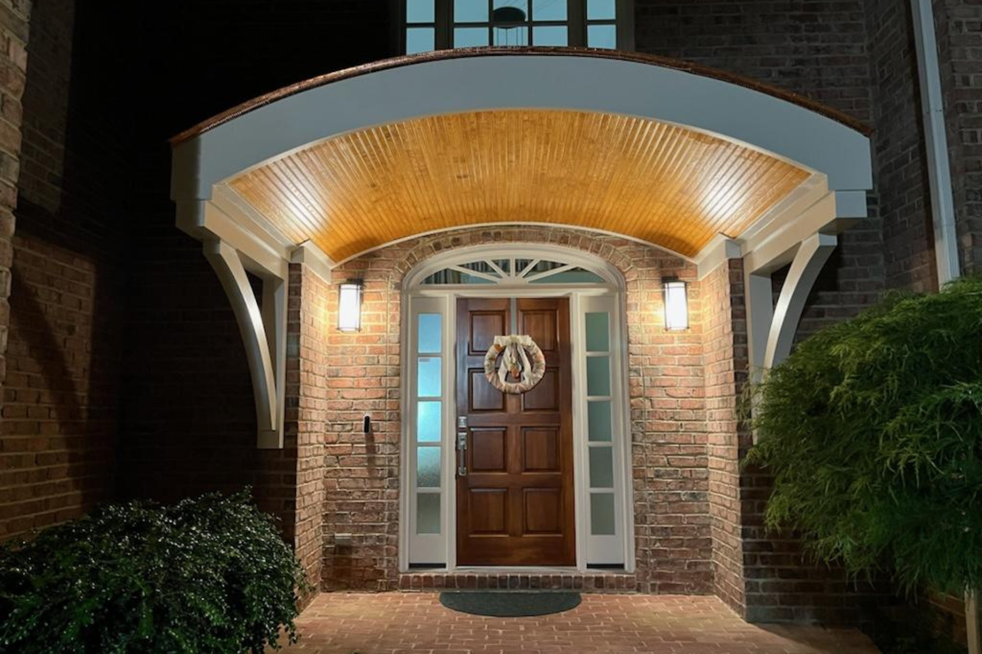 A lit wooden front door with a wreath under an arched portico, framed by brick walls and greenery at night.