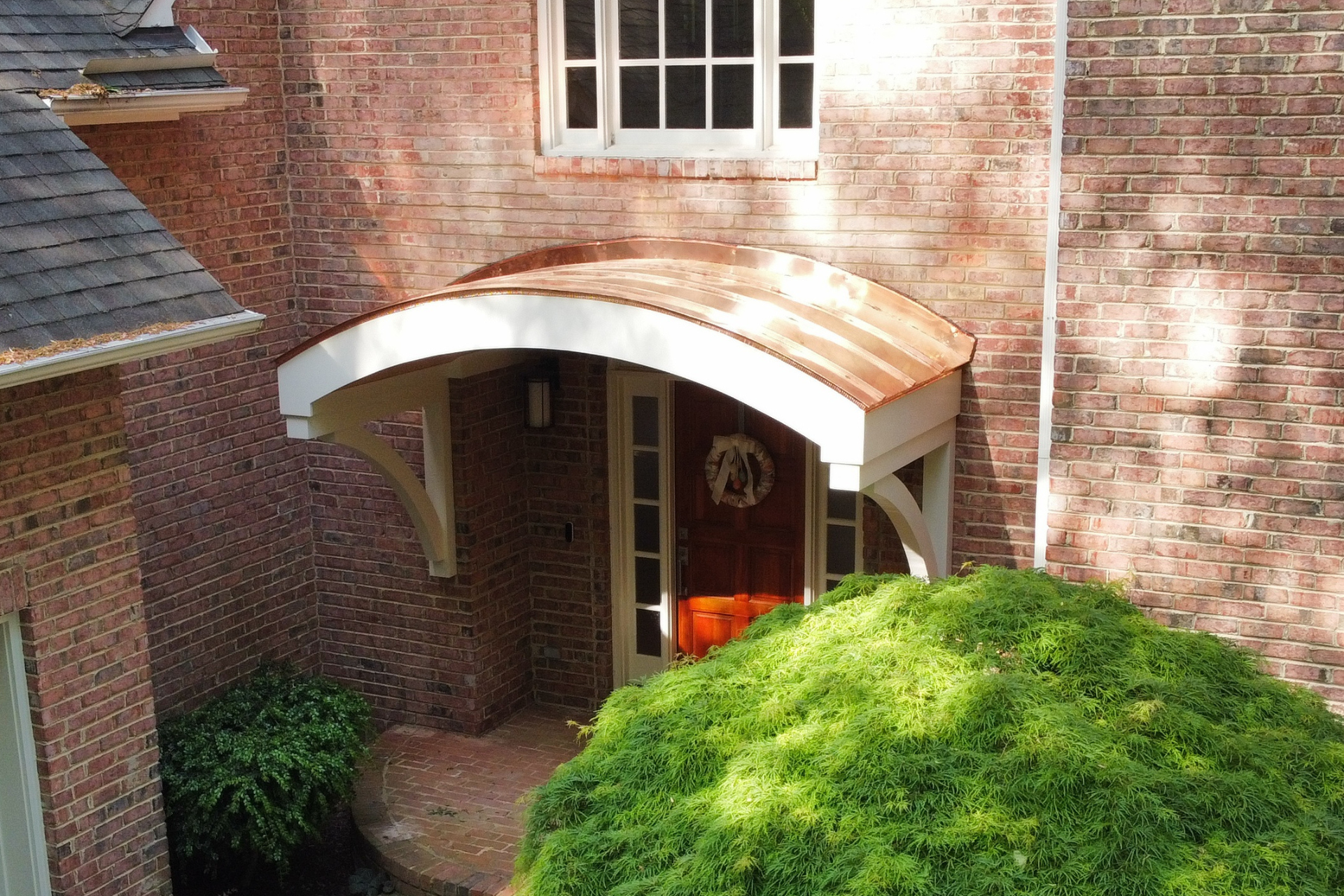 A brick house exterior featuring a curved copper-roofed entryway porch over a wooden door, framed by green landscaping.