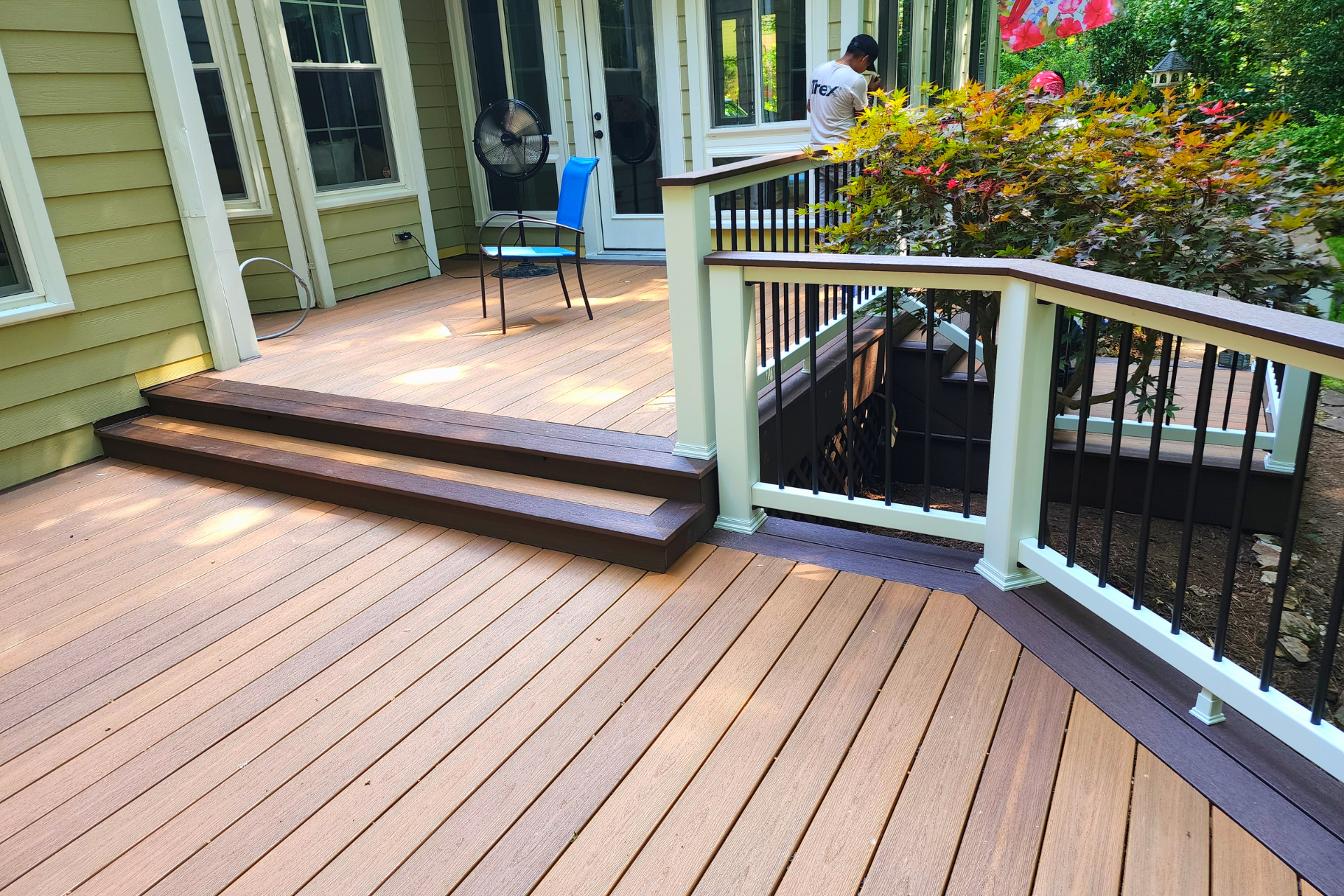 A light brown wooden deck with two steps, a white railing with black spindles, and a chair near a house entrance.