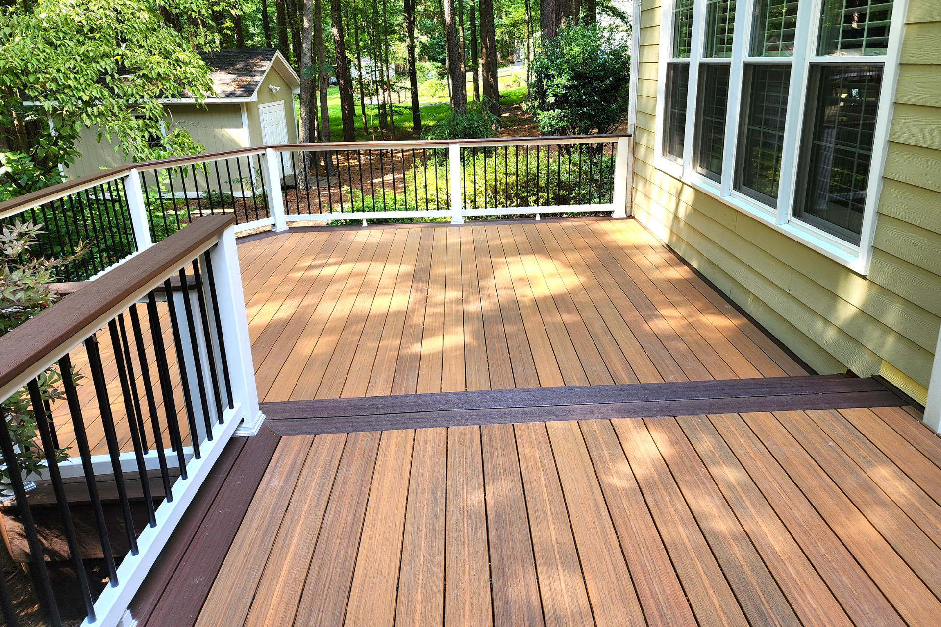 A wooden deck with a contrasting dark border and black metal railings with white posts, overlooking a wooded yard.