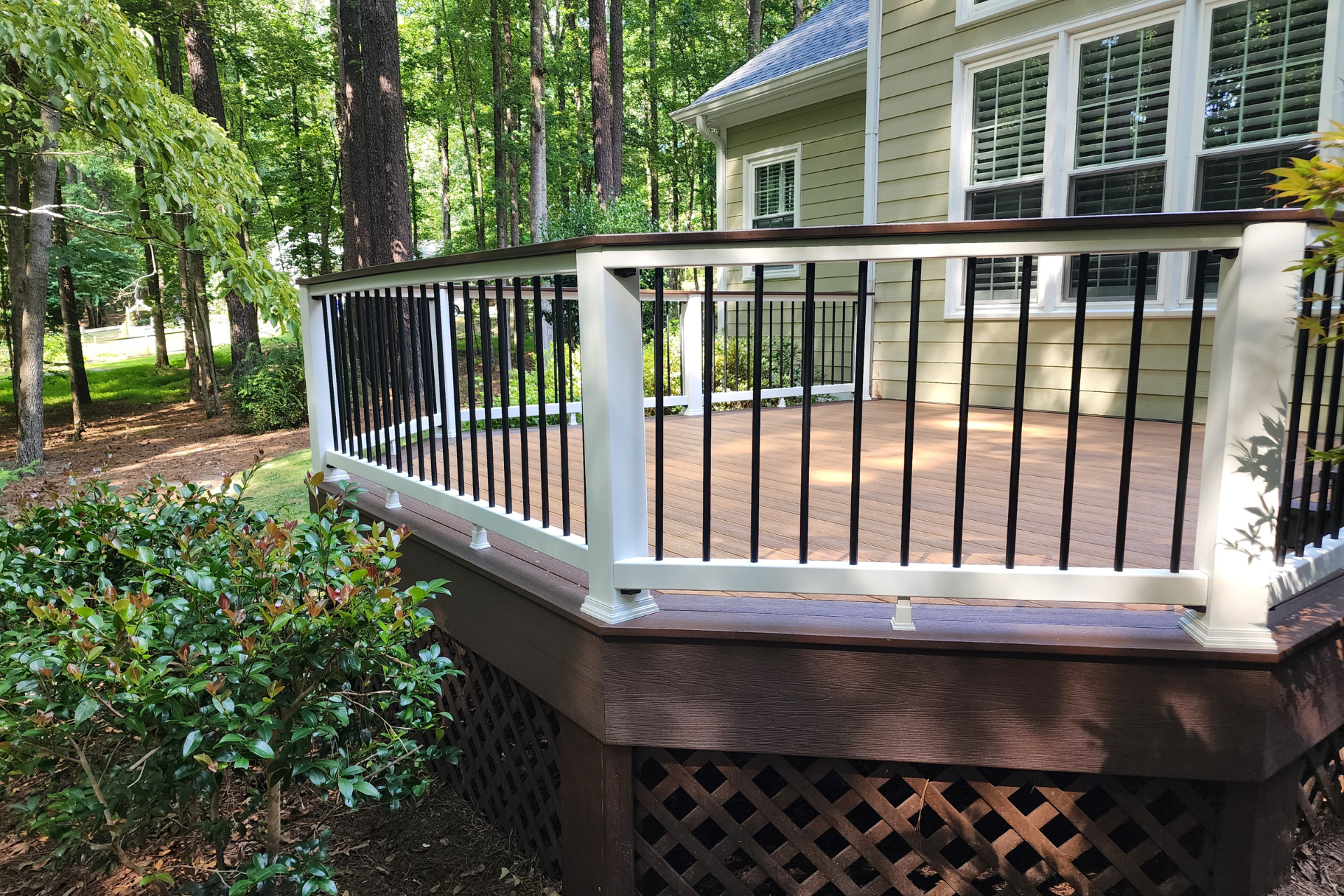 A raised deck with white railings, black balusters, and lattice skirting, situated next to a house by a wooded area.