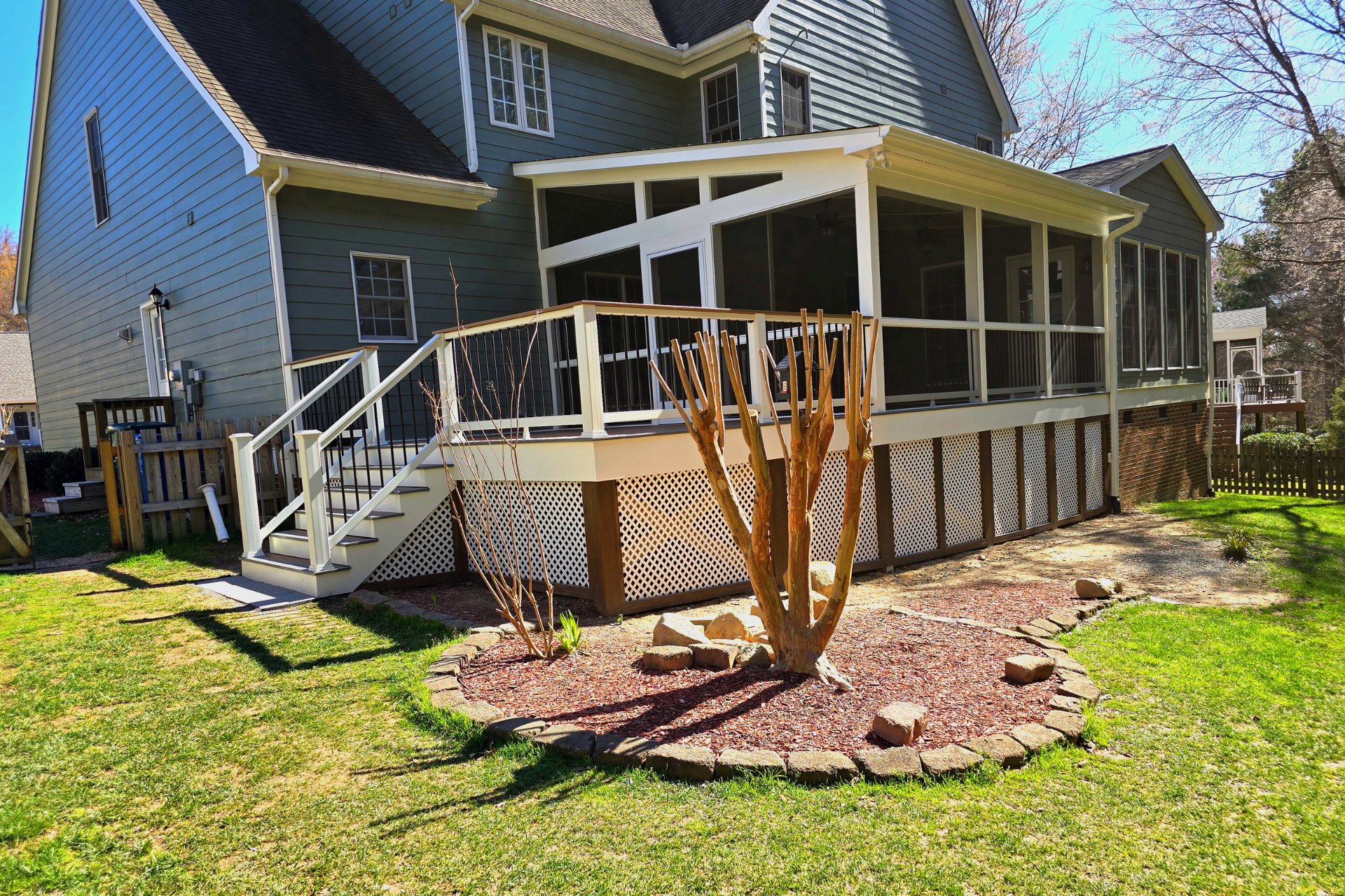 A gray-sided house with a white screened-in porch and a wooden deck with stairs over a landscaped yard with a small tree.