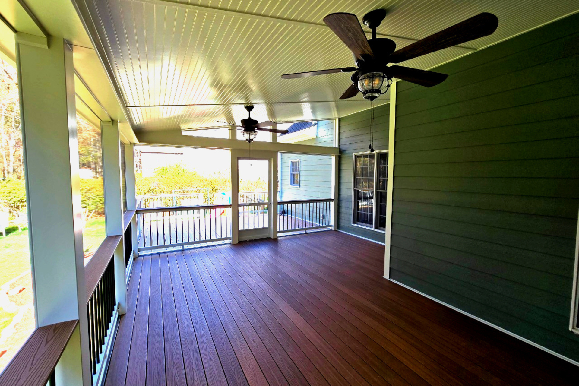 A covered porch with wood-look flooring, dark green siding, white railings, and two ceiling fans.