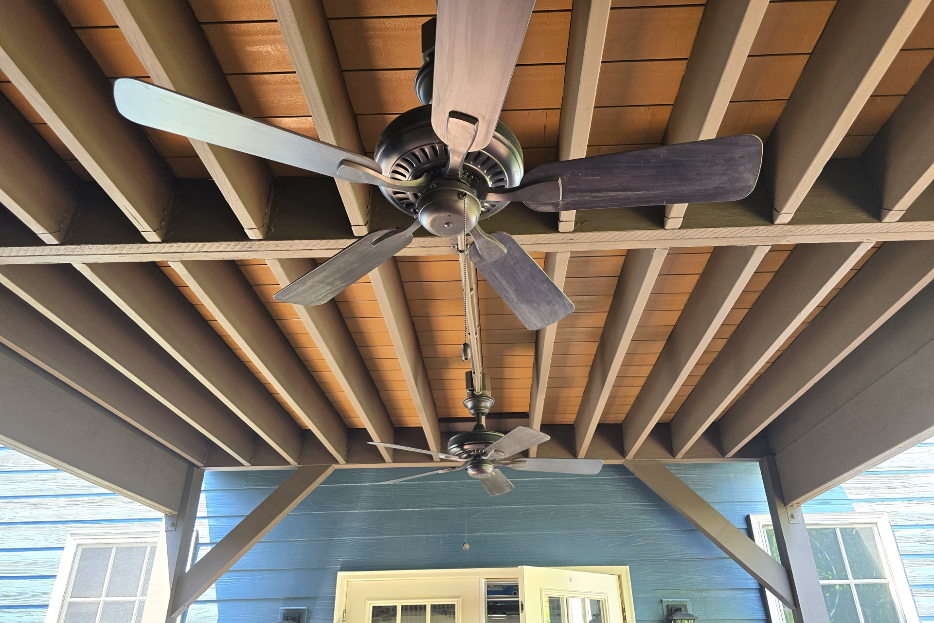 Two dark-colored ceiling fans suspended from the exposed wooden ceiling beams of an outdoor covered porch.