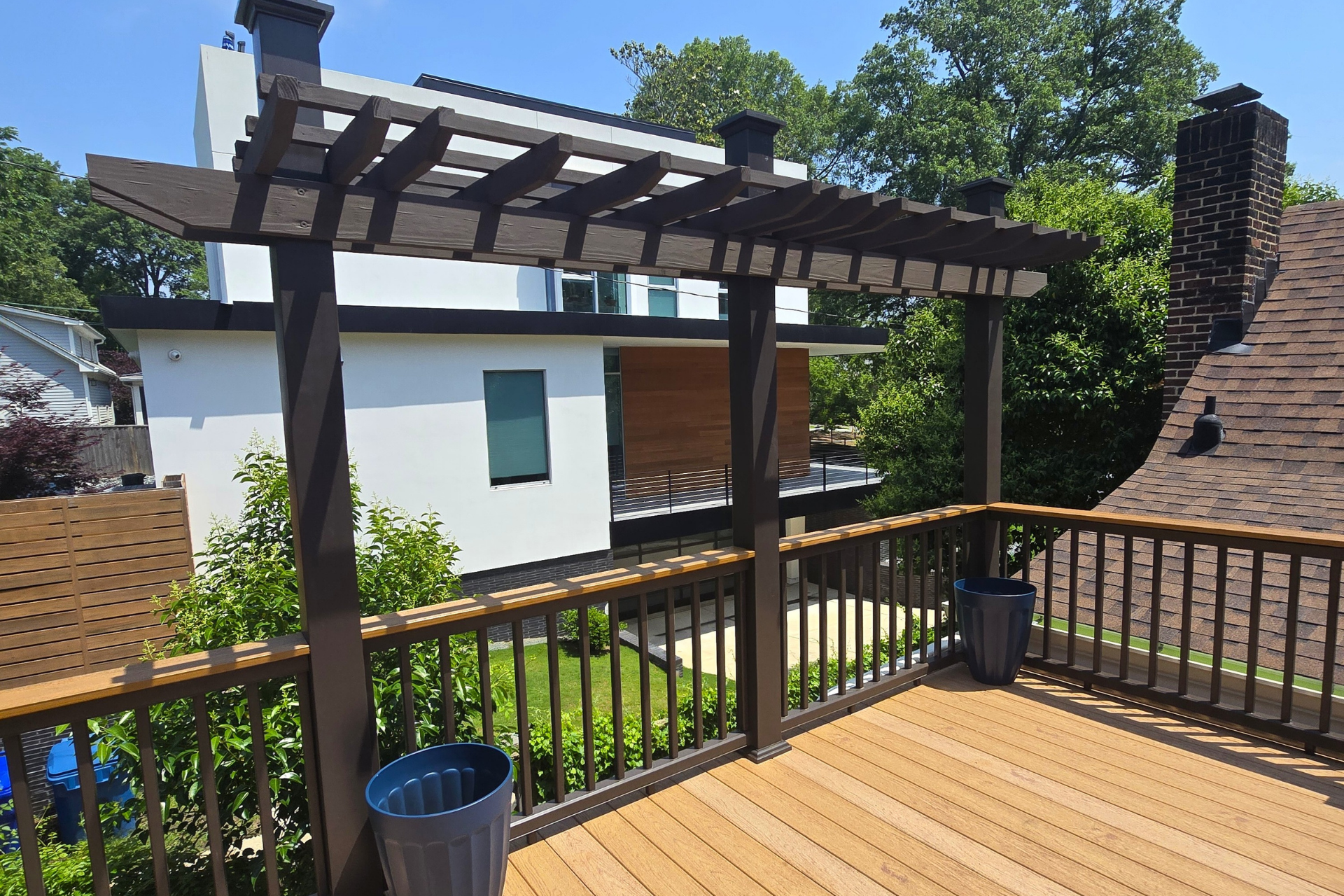 A wooden deck with a dark brown pergola, railing, and matching pots overlooking a backyard and a modern house.