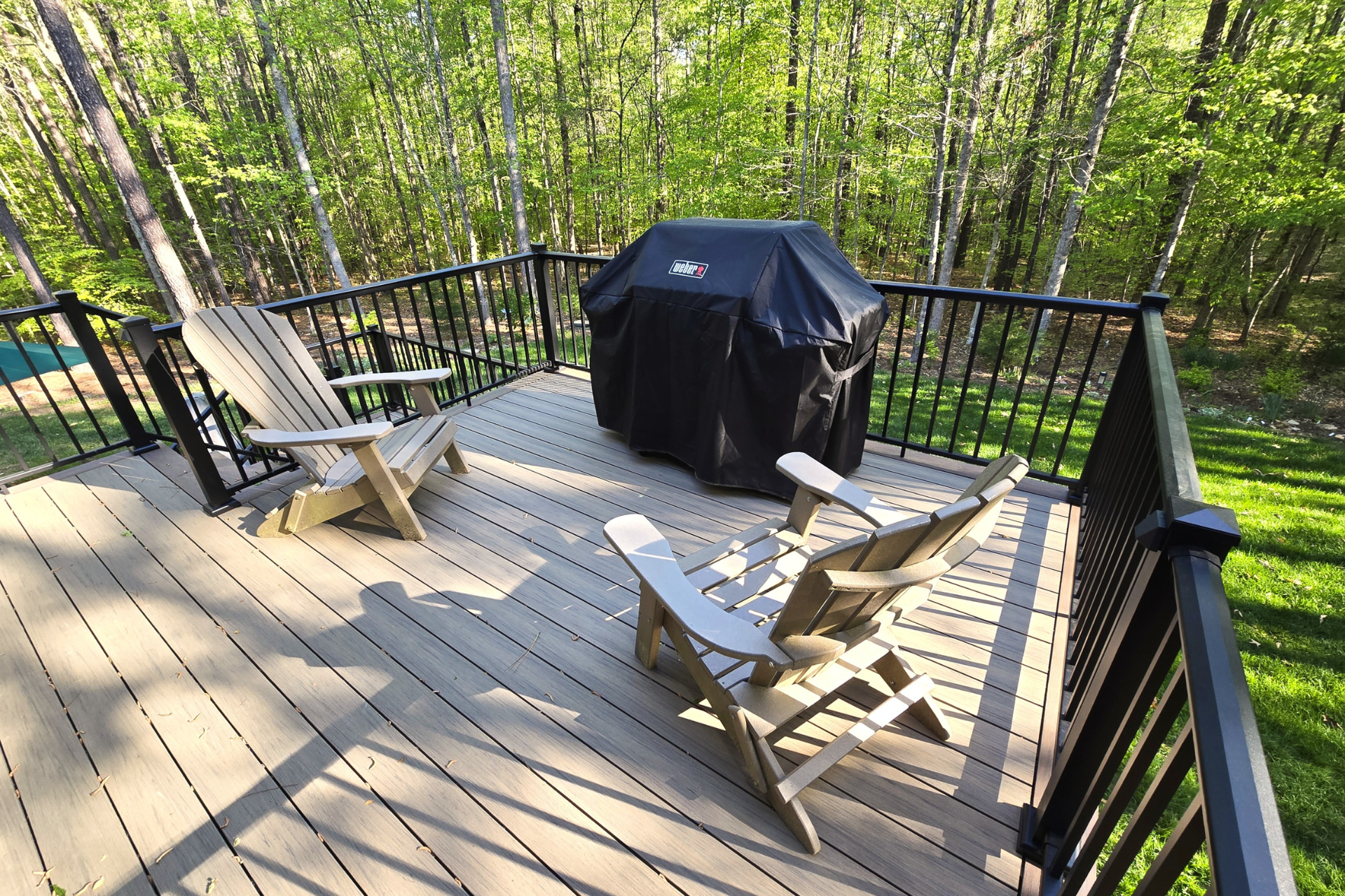 Two wooden Adirondack chairs and a covered grill on a gray deck railing, overlooking a lush green forest.