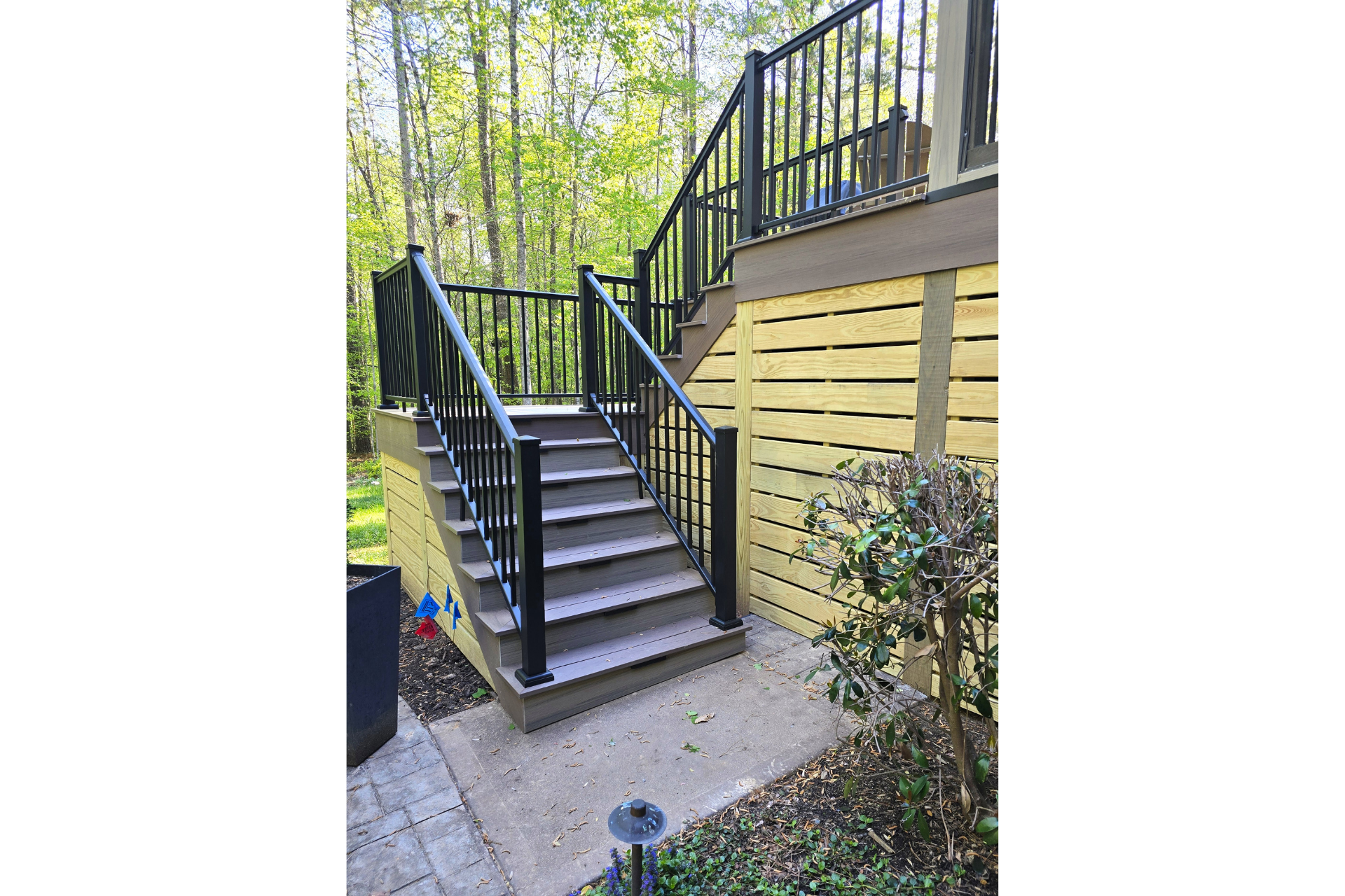 Outdoor wooden stairs with black railings lead to a raised deck against a yellow-sided house with a surrounding garden.