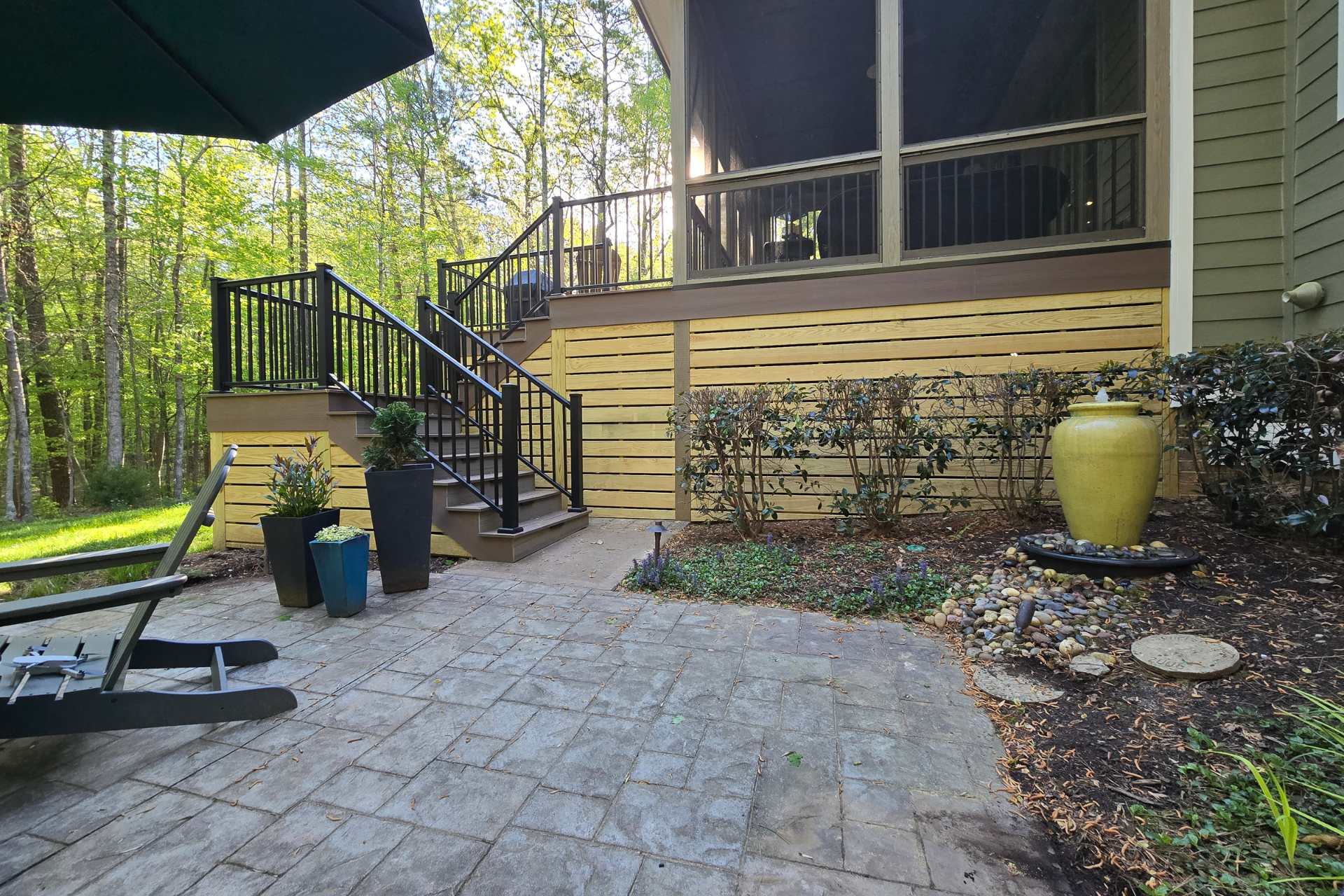 A stone patio with a lounge chair, potted plants, a water feature, and stairs leading to a deck with horizontal siding.