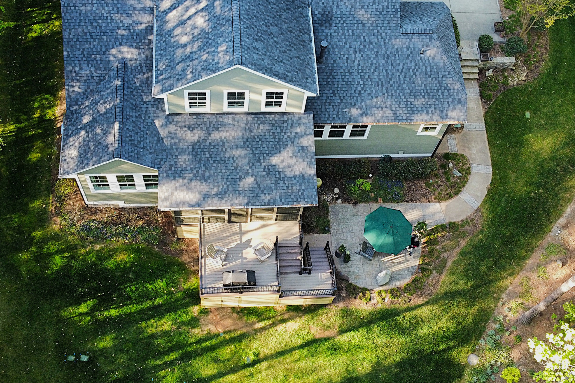 Aerial view of a house with a gray roof, wooden deck, green patio umbrella, and stone walkway on a lush, grassy lawn.