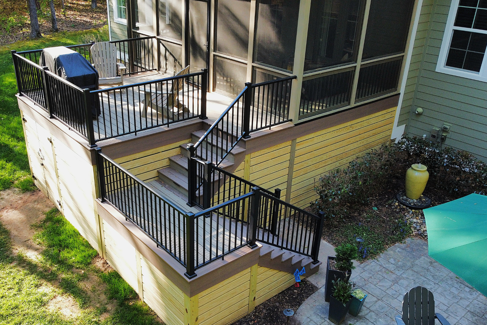 A multi-level yellow deck with black railings and stairs leading to a stone patio with a green umbrella.