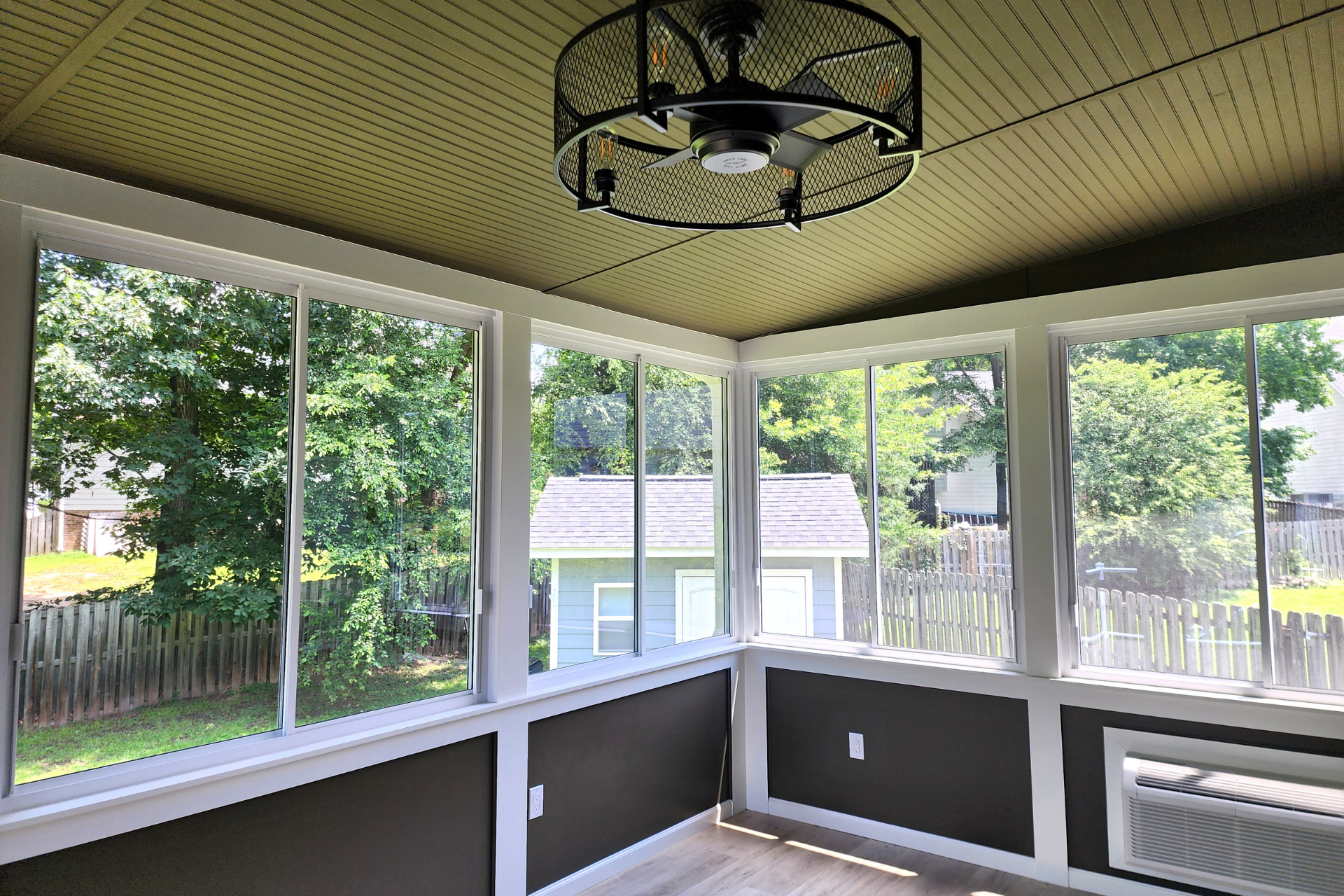 A sunroom with large windows, dark grey lower walls, a wood-paneled ceiling, and a black cage-style ceiling fan.