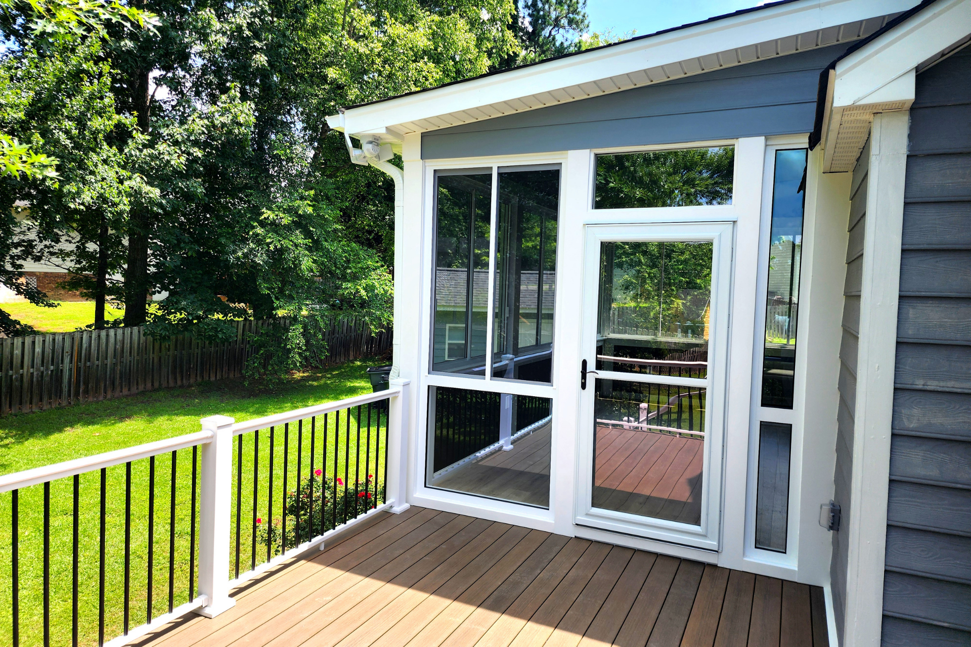 A wood-toned deck with a white railing overlooks a green backyard next to a sunroom with large white-framed windows.