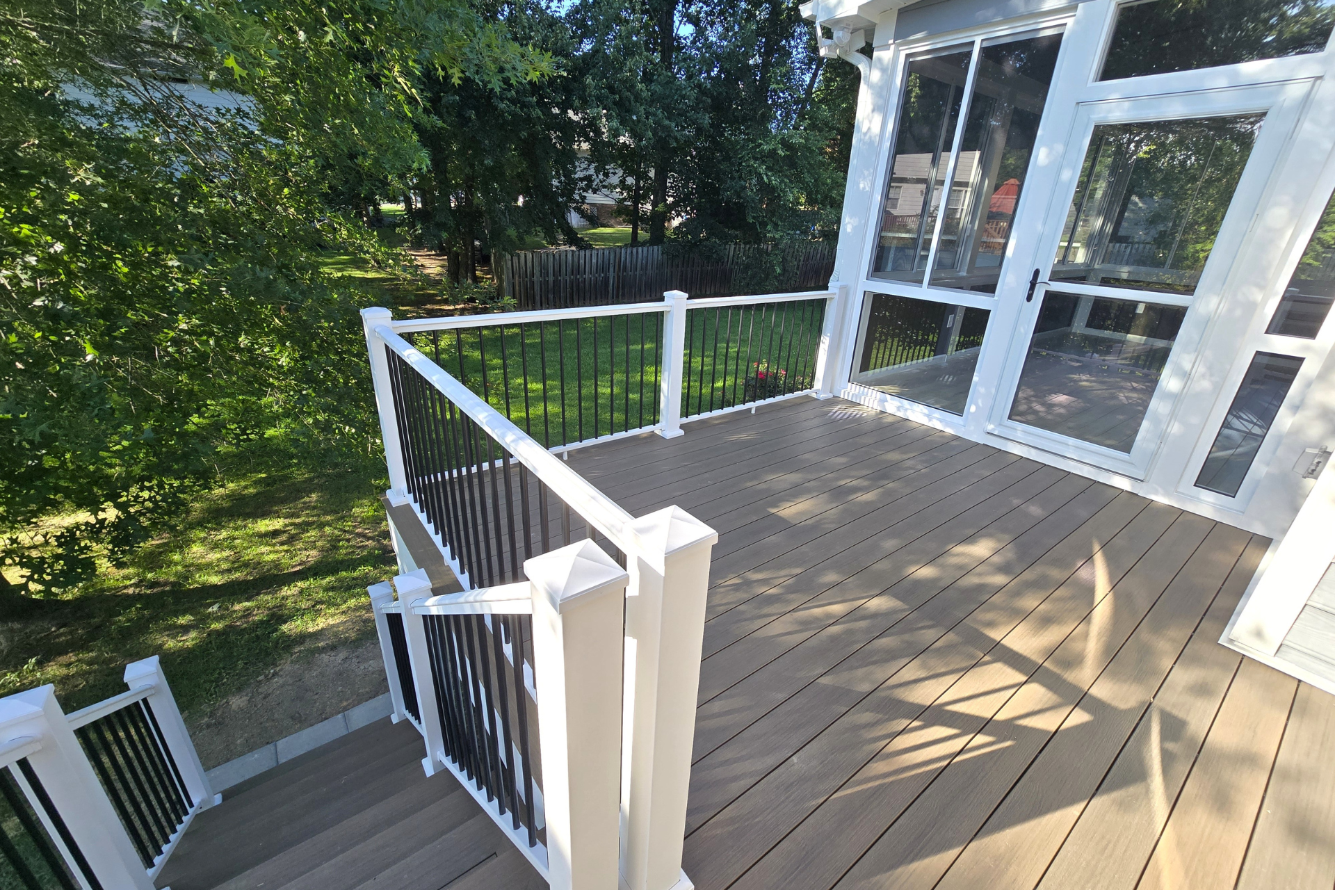 A raised deck with light brown composite flooring, white posts, black railings, and stairs leading to a grassy yard.